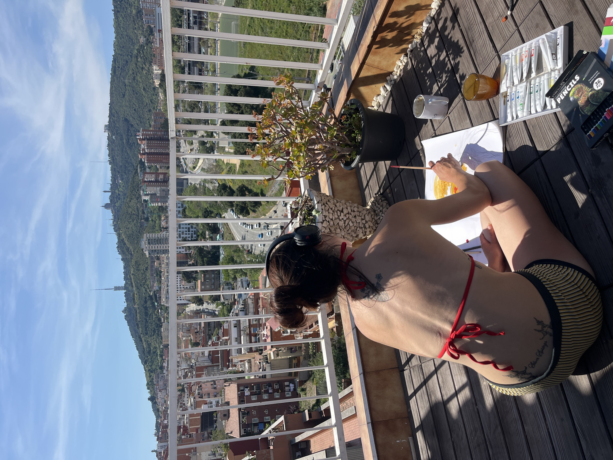 A woman in a red bikini top and striped swimsuit bottoms sits on a wooden balcony, painting on paper. The balcony overlooks a cityscape with a tall mountain in the background, characteristic of Barcelona, Spain. The scene includes painting supplies: a set of tubes, a box of colored pencils, water cups, and a brush. There is also a potted plant on the balcony. She is wearing headphones. Tattoos are visible on her skin.