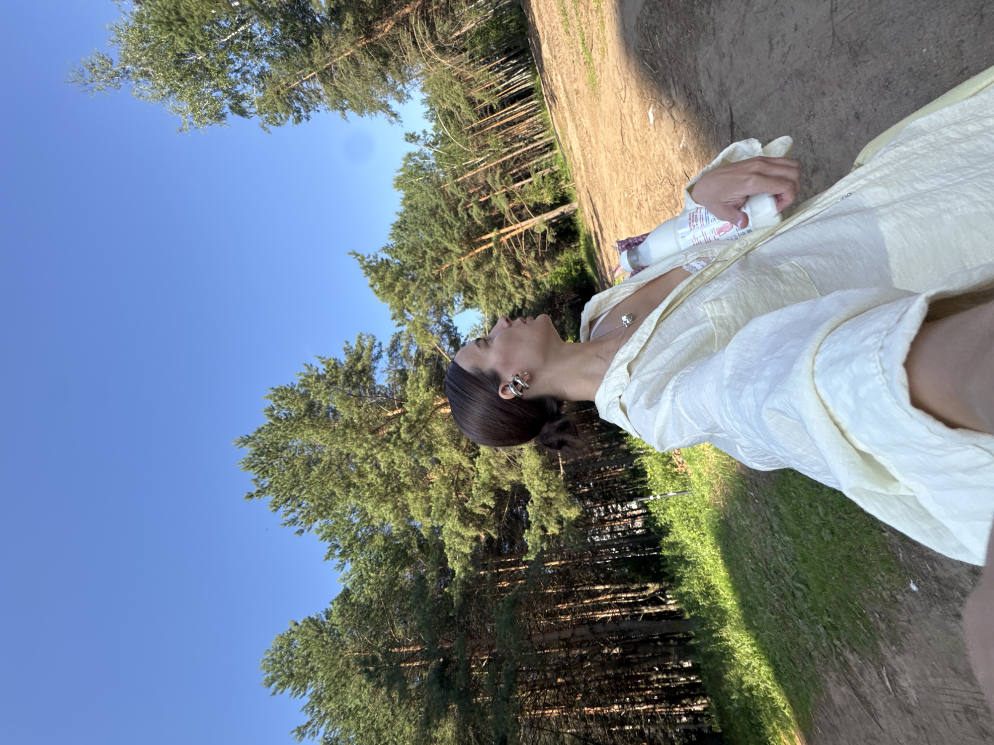 In a natural environment with tall pine trees and a clear blue sky, a woman is holding a bottle of liquid. The woman, with her hair in a bun and wearing a light-colored jacket, is looking to her right. She has a necklace with a heart-shaped pendant and earrings. The ground is a mix of dirt and grass. The overall scene suggests an outdoor activity.