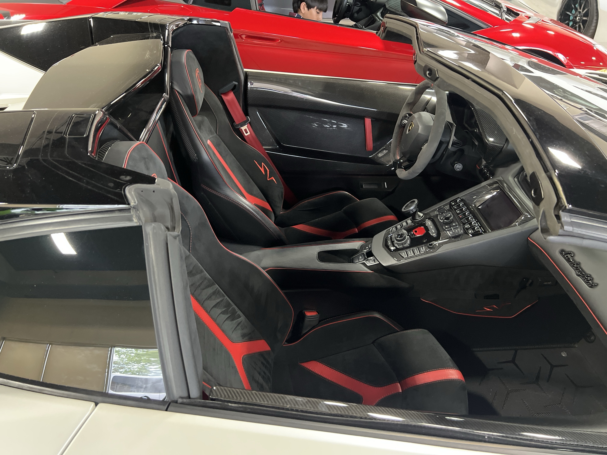 The image shows the interior of a black and white Lamborghini Aventador SVJ roadster. The car features black suede seats with red accents, and a gray dashboard. In the background, there's a red Lamborghini with a person sitting in the driver's seat. Another part of the red car is visible in the upper right corner of the frame. The location appears to be indoors, possibly a showroom or garage. The city and country are not visible in the image.