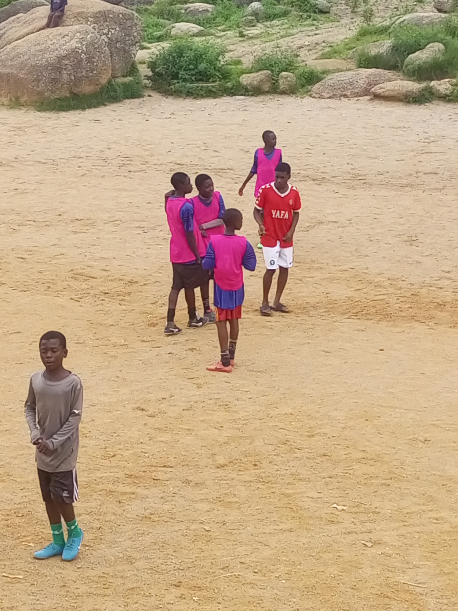 The image depicts a group of young individuals, likely boys, gathered on a sandy field in what appears to be an outdoor recreational area. The background features large rocks and sparse vegetation, suggesting a dry, possibly arid environment. A few individuals are wearing athletic attire, including t-shirts, shorts, and soccer cleats, indicating a sporting activity, possibly a soccer practice or game. The overall atmosphere seems casual and informal. This scene is located in Jos, Nigeria, a city known for its rocky terrain and plateau landscape.