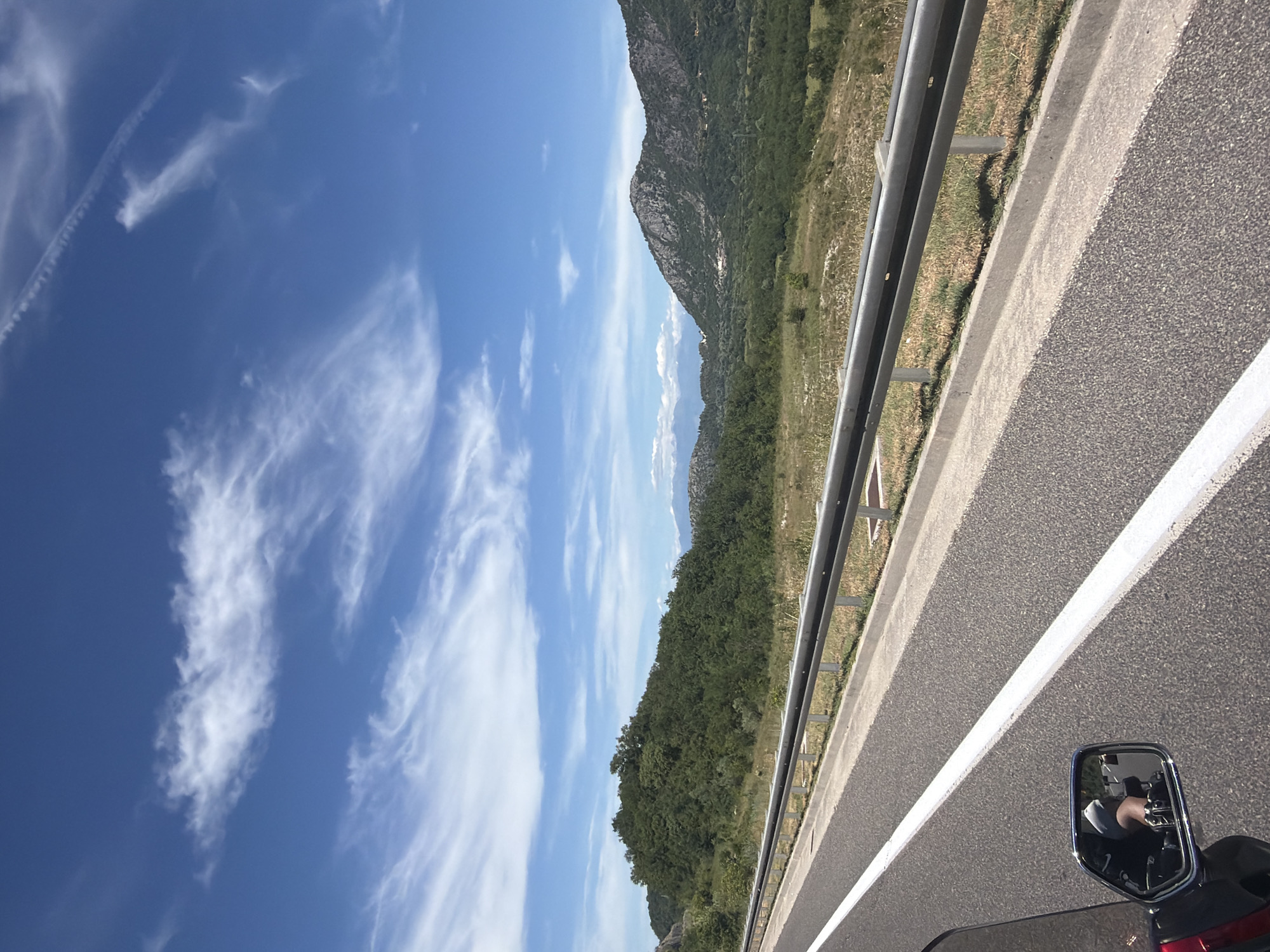 The image depicts a view from a vehicle traveling on a highway. In the foreground, a paved road with a white dividing line is visible, bordered by a metal guardrail and a concrete curb. Beyond the road, a lush green landscape unfolds, characterized by rolling hills covered in dense vegetation and trees. In the background, a mountain range with rocky peaks is discernible against a clear blue sky dotted with wispy white clouds. The scene appears to be set in a rural, mountainous region, possibly near Sotonići, Montenegro, given the mountainous terrain and vegetation. A side mirror of the vehicle reflects a person's legs and feet, wearing shorts and sneakers, indicating the perspective of someone riding in the vehicle. The overall atmosphere is one of travel through a scenic, natural environment on a sunny day.