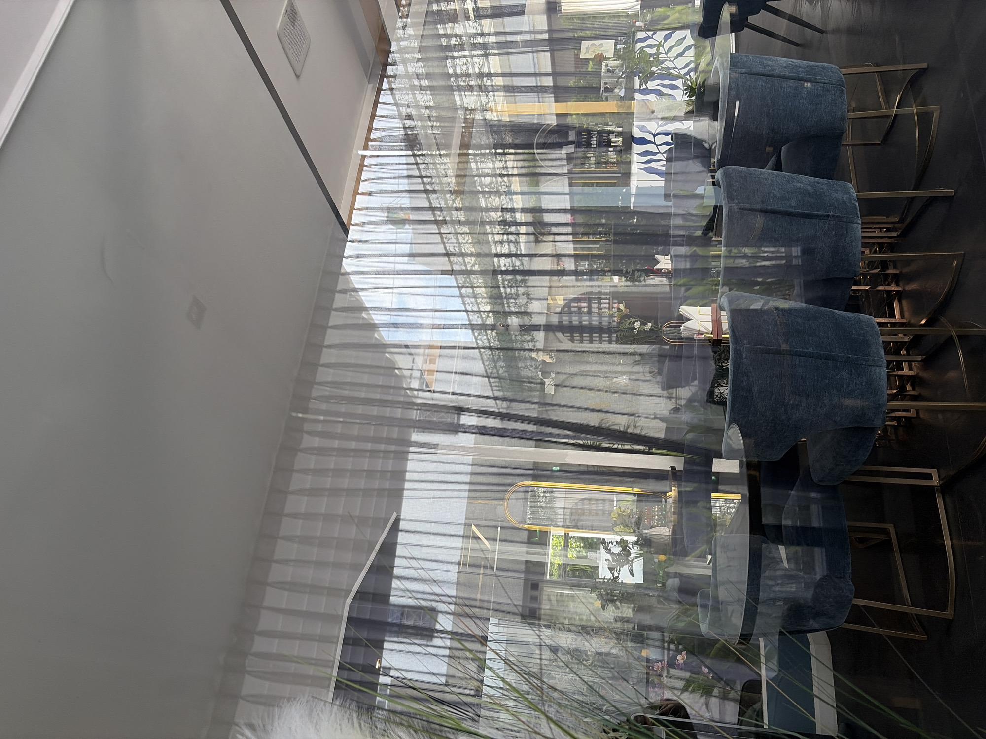 The image shows the interior of a restaurant, seen through a glass partition or window, with reflections obscuring some details. In the foreground, there are several blue velvet chairs with gold metal legs arranged around dark, round tables. Some of the tables appear to be made of glass. To the left, a tall, feathery plant is visible.
Behind the chairs and tables, the restaurant's interior is visible through sheer white and dark vertical curtains, and reflections. There are more tables and chairs, as well as decorative elements like plants, bottles on shelves, and a large gold-framed mirror with an oval shape. The architecture suggests a modern design with large windows and a high ceiling. Natural light filters into the space, creating a bright atmosphere. The overall impression is of a well-lit, contemporary dining establishment.