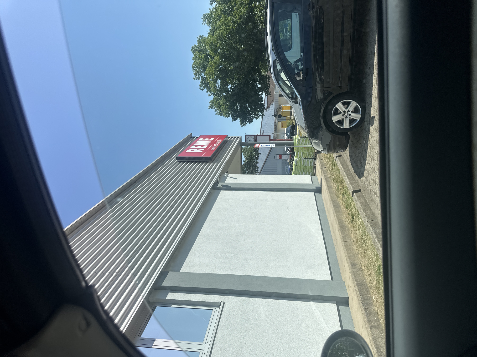 An exterior, eye-level, medium shot captures a modern building with a grey textured facade and a corrugated metal upper section, on a sunny day. A red sign with white lettering, reading "REWE Familie Schölche", is affixed to the building's facade. To the right of the building, a black car is parked in a lot. The car is a Volkswagen Polo, seen from its right side. Behind the car, a large green tree with dense foliage dominates the background. Further back, solar panels are visible on a roof. The scene appears to be a commercial area, possibly a supermarket parking lot. The image is framed from within a vehicle, suggesting the photographer is seated inside.