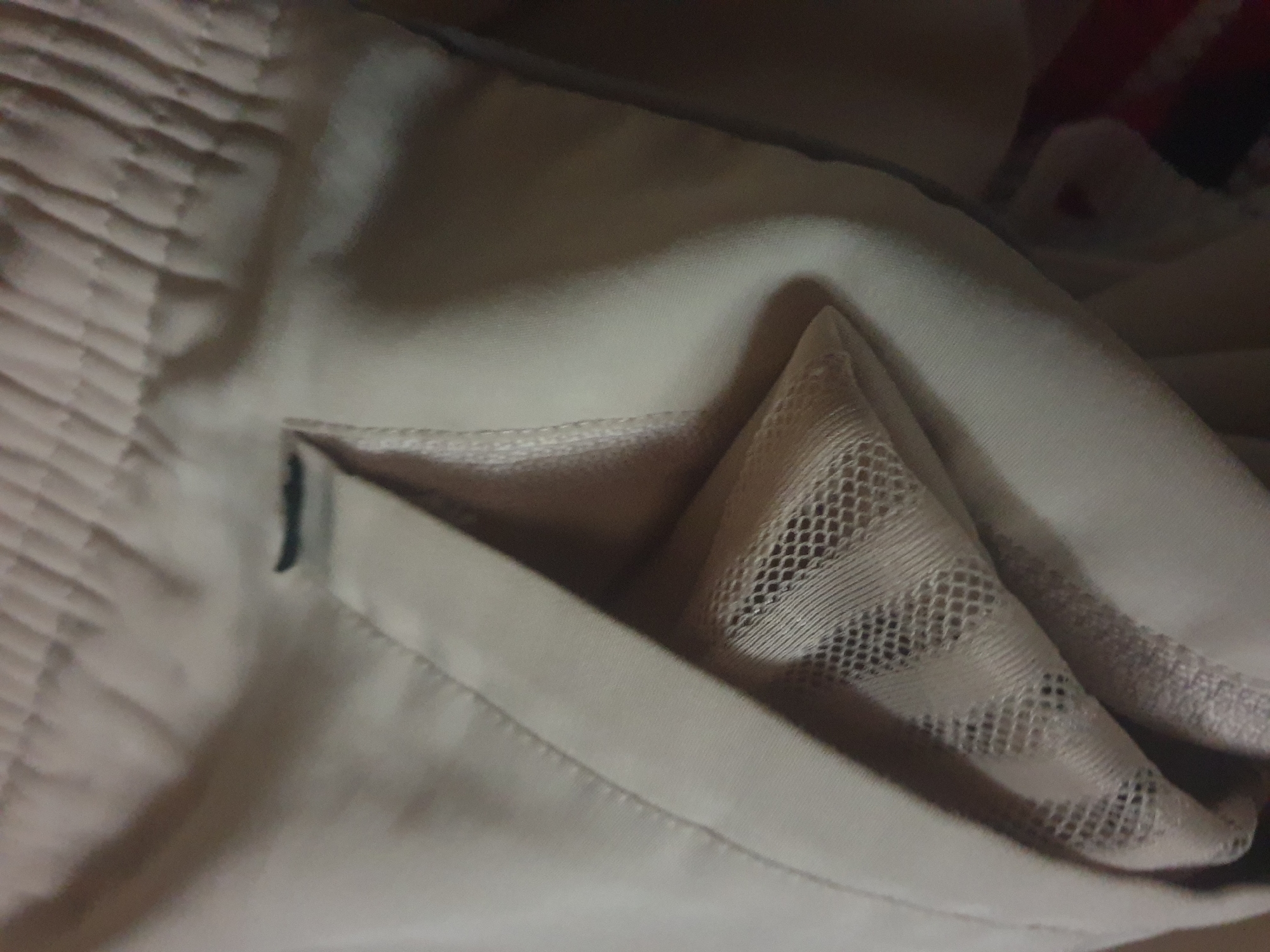 The image shows a close-up view of a light-colored fabric with a mesh material partially visible within a pocket. The fabric appears to be part of clothing, possibly shorts or pants, due to the elastic waistband visible at the top of the image. There is a dark slit on the waistband, which could be a buttonhole or a decorative element. The lighting is dim, casting shadows that obscure some of the details. There are no people, animals, or visible location indicators in the image. The context of the city or any specific location is not discernible.