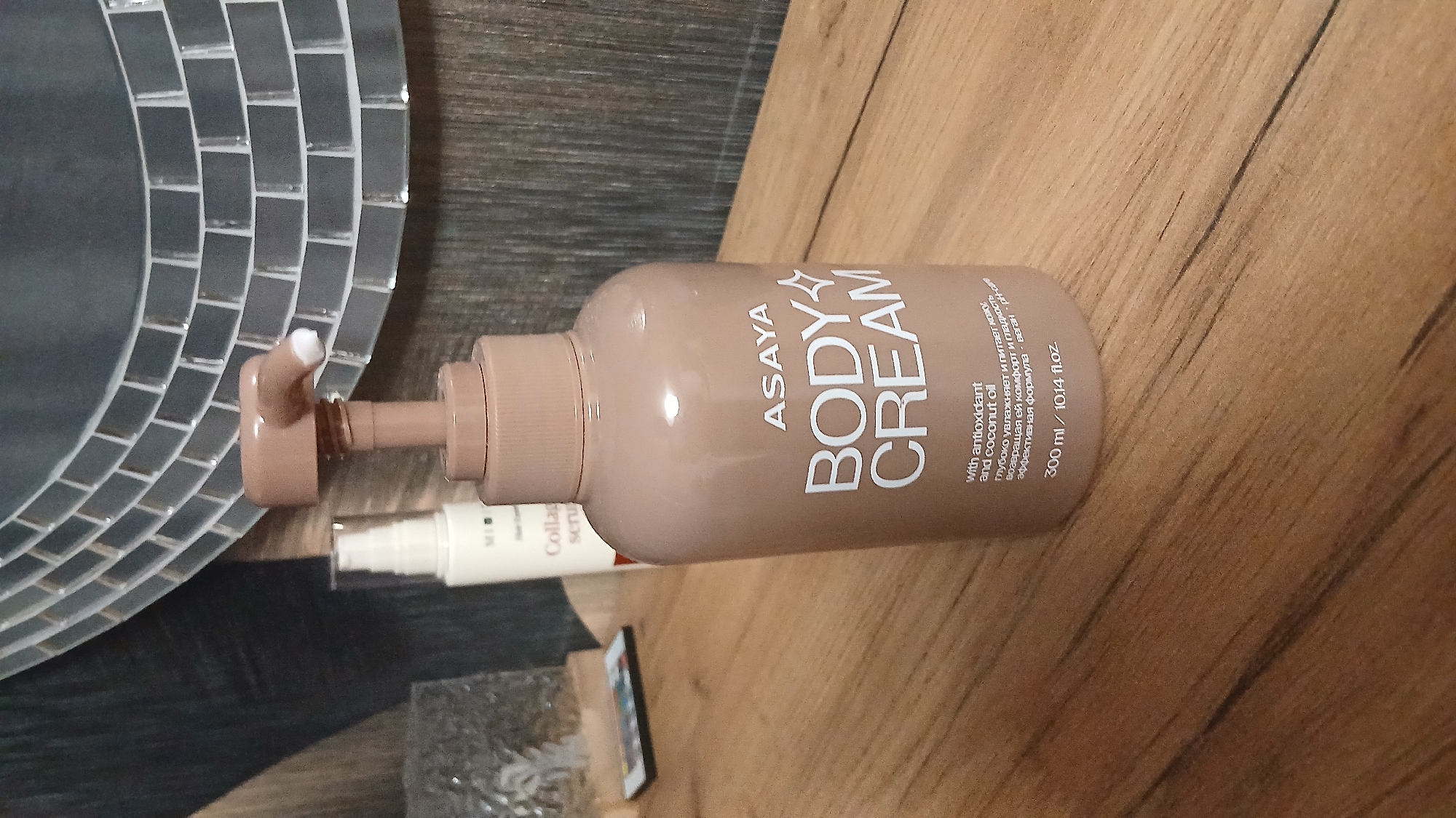 An overhead shot shows a light brown pump bottle of ASAYA BODY CREAM standing on a wooden surface. The bottle has white text on it, and the pump mechanism is also light brown. A smaller white bottle with a clear cap is visible behind it. In the background, a decorative mirror with a mosaic of grey tiles is visible. The overall scene is indoors, with a neutral, uncluttered aesthetic.