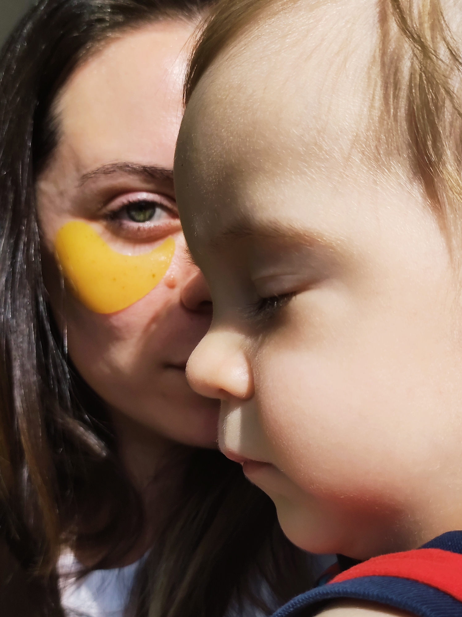 A close-up shot depicts a woman and a child. The woman is partially visible behind the child. She has brown hair and green eyes. There is a yellow under-eye patch on her face. The child, in the foreground, has light blonde hair and appears to be looking down. The child is wearing a dark blue shirt with red stripes. The background is blurry. The location is Chaikovsky, Russia.