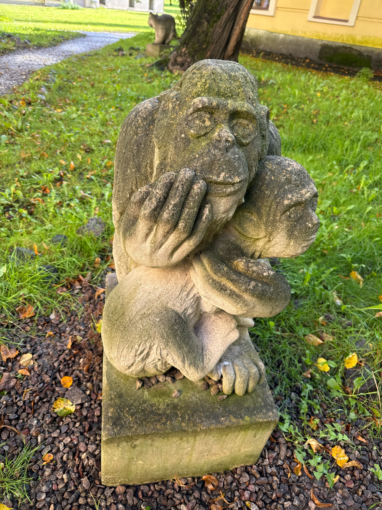 A stone sculpture of two monkeys is situated on a stone base in a grassy area. The larger monkey, in the foreground, is embracing a smaller monkey. Both figures are covered in moss and lichen, indicating age and exposure to the elements. In the background, a path leads away from the sculpture, with a larger tree to the right of the monkeys. Beyond the path, a building with yellow walls and rectangular windows is visible, suggesting an urban or park-like setting. Fallen leaves are scattered on the grass and ground around the sculpture. The overall impression is of an outdoor sculpture in a slightly neglected but natural environment. The location is not explicitly identifiable as Veliky Novgorod, Russia, based on the visual information provided.