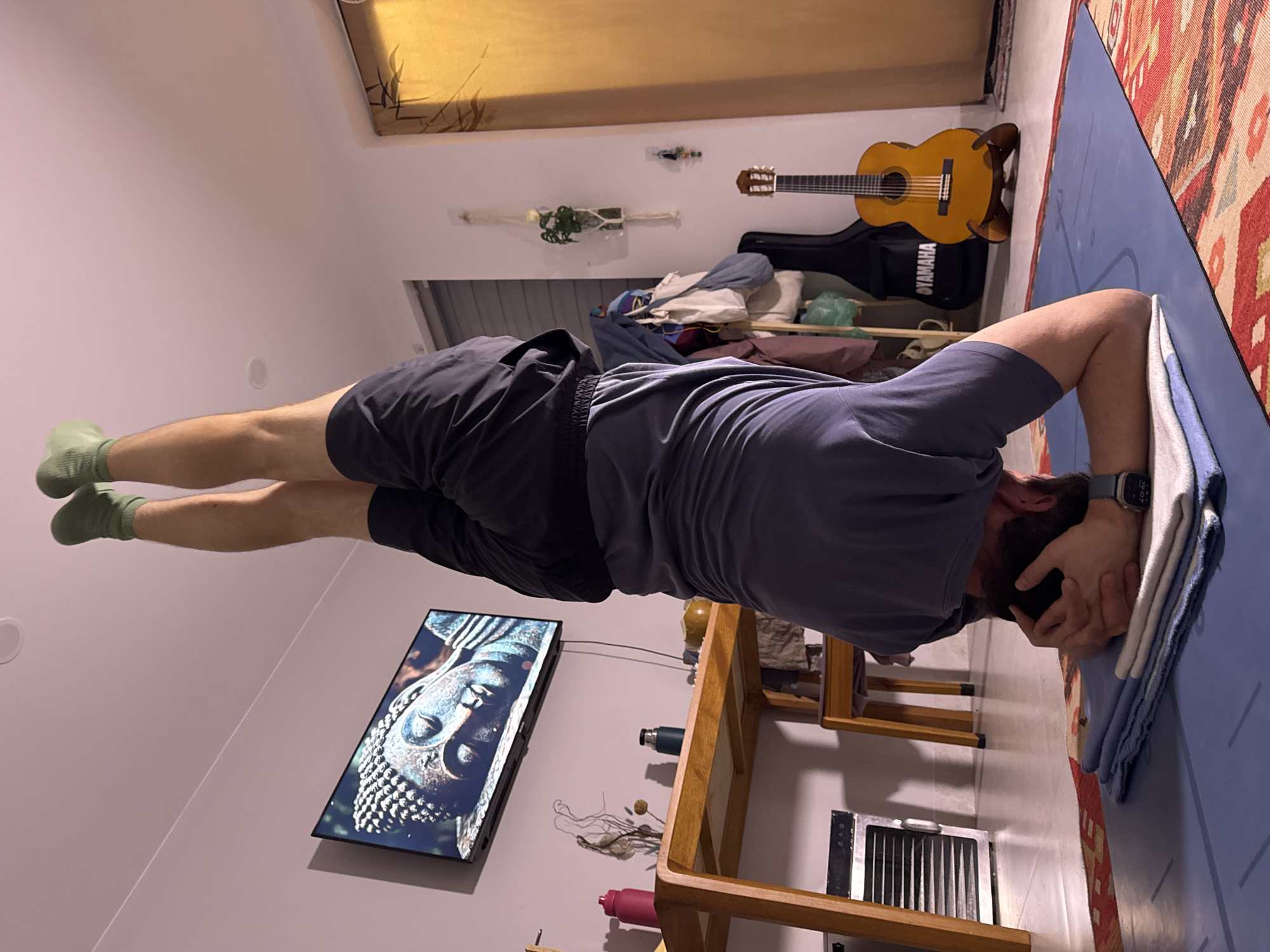 A man is performing a headstand on a blue yoga mat. His head is resting on a stack of white and blue towels. He is wearing a gray t-shirt, black shorts, and green socks. A guitar and guitar case are visible in the background, along with a television screen displaying an image of Buddha. The location is likely inside a home. I have no way of knowing if this is in the city of Lâm Viên - Đà Lạt, Vietnam.