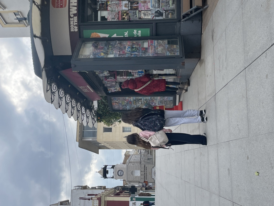 This image captures a street scene in Mérida, Spain, centered around a newsstand on a cloudy day. In the foreground, a newsstand with a distinctive striped awning, supported by ornate wrought iron, is prominently displayed. Its shelves are filled with various magazines and newspapers, some featuring titles like "HISTORIA." The side of the newsstand displays advertising text, including fragmented phrases such as "BURG KIP" (likely "BURGER KING") and "HOME OF THE WH" (likely "HOME OF THE WHOPPER"). The word "HOY" (Spanish for "Today") is also visible on its facade.
Several individuals are engaged with the newsstand. One person, wearing a red coat, is actively browsing the displayed items. Nearby, two other younger individuals with long hair are also looking towards the stand; one wears a floral patterned backpack, and the other a light-colored backpack. A fourth person is partially visible further down the street.
The street is paved with light-colored tiles, indicating a pedestrian area. In the background, an urban landscape unfolds with various buildings. A notable architectural feature is a clock tower with a dome, part of a larger building that bears text identifying it as "OFICINA DE TURISMO" (Tourist Office) and "Ayuntamiento de Mérida" (City Hall of Mérida), confirming the location. The sky is overcast, suggesting a daytime scene without direct sunlight. Overhead wires stretch across the street.