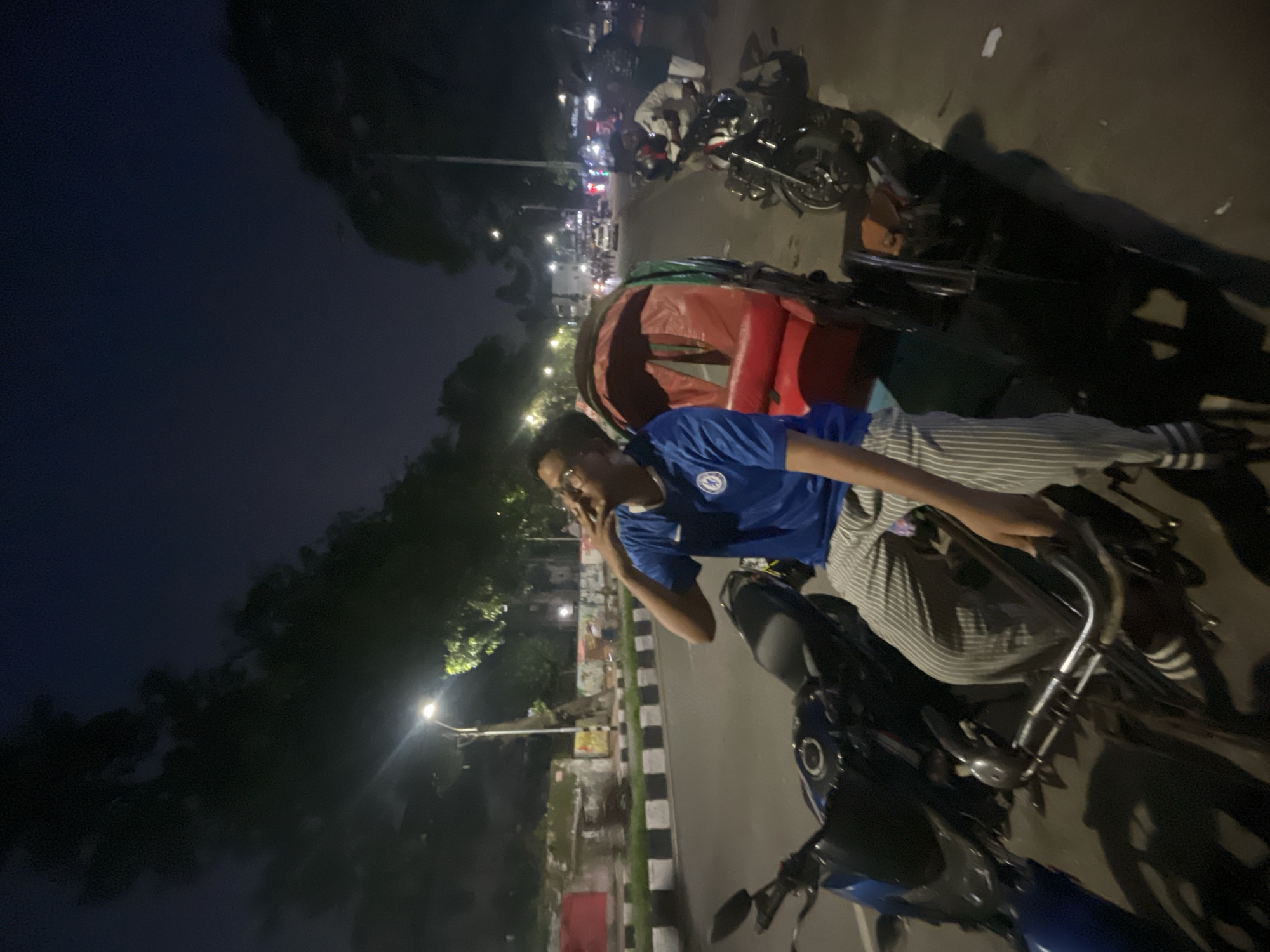 A man wearing a blue shirt and striped pants sits on a motorcycle in Dhaka, Bangladesh, and is on the phone. The photo was taken at night. Parked motorcycles and cycle rickshaws are nearby. Street lights illuminate the road and the surrounding area.