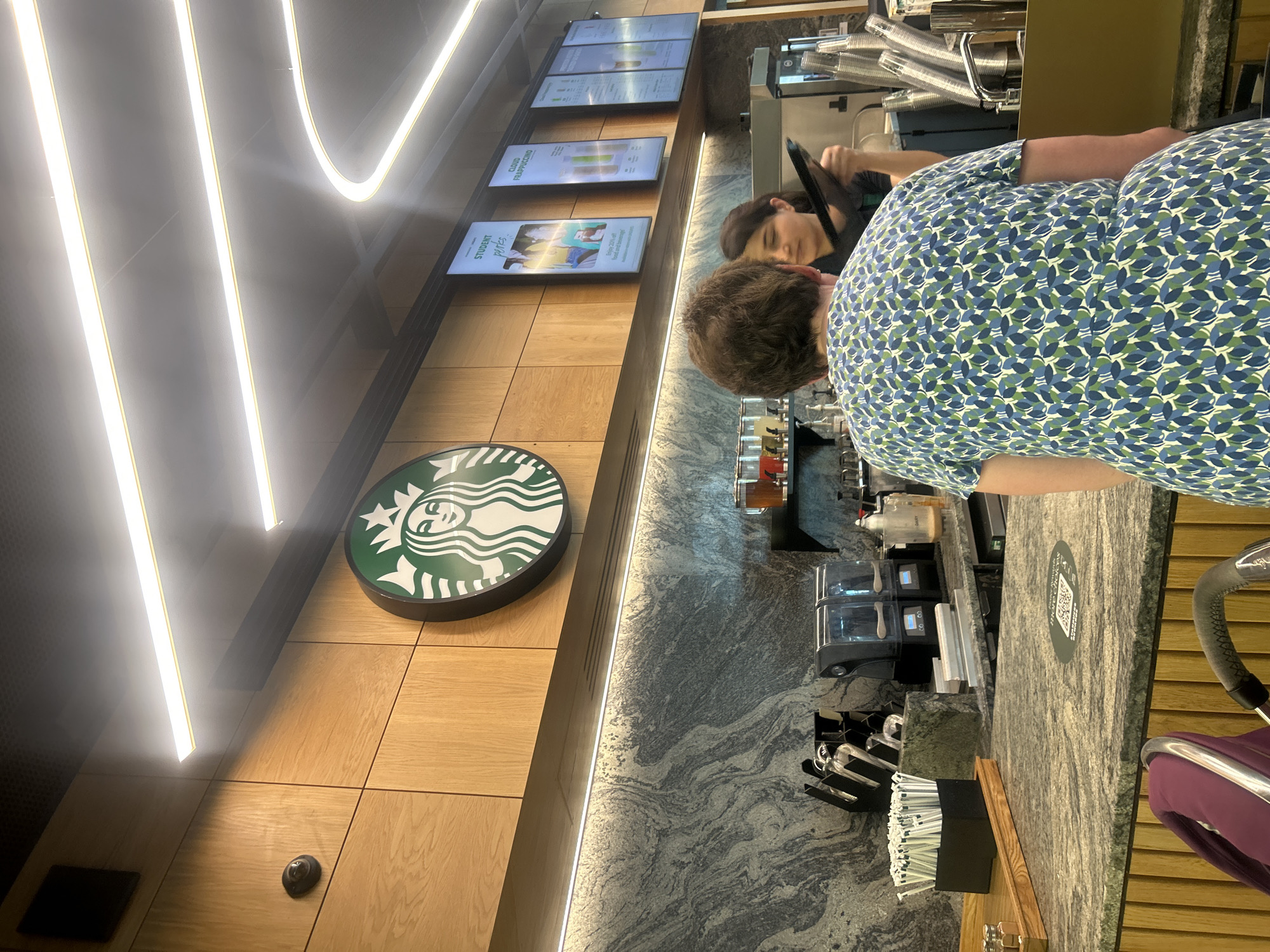 The image shows the interior of a Starbucks store in London, United Kingdom. The Starbucks logo is prominently displayed on a wood-paneled wall near the counter. Overhead menus display drink options. A customer wearing a blue and green patterned shirt is at the counter, where a Starbucks employee wearing a black uniform is working. A stroller is near the counter. The counter is made of gray stone with swirling patterns. The overall lighting is bright, with LED strip lighting on the ceiling and walls. Utensils, machines, and various containers are visible on the counter behind the employee.