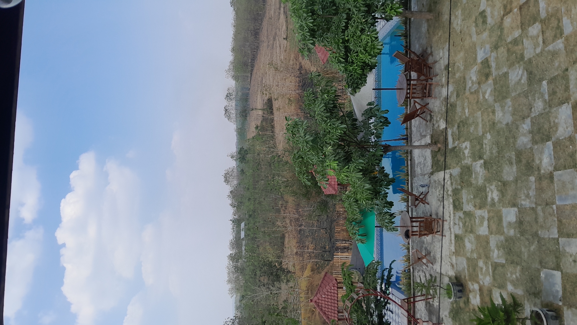 The image shows an outdoor scene with a swimming pool and surrounding landscape. In the foreground, there is a paved area with several sets of wooden tables and chairs. Some potted plants are also visible. A swimming pool with blue water is situated behind the paved area, with a green umbrella near one end. In the background, there are trees and rolling hills, suggesting a rural or semi-rural environment. The sky is partly cloudy and a light haze is present in the distance. The scene appears to be in a resort or recreational area. No people are visible in the image. The location is identified as Giyanti, Indonesia.