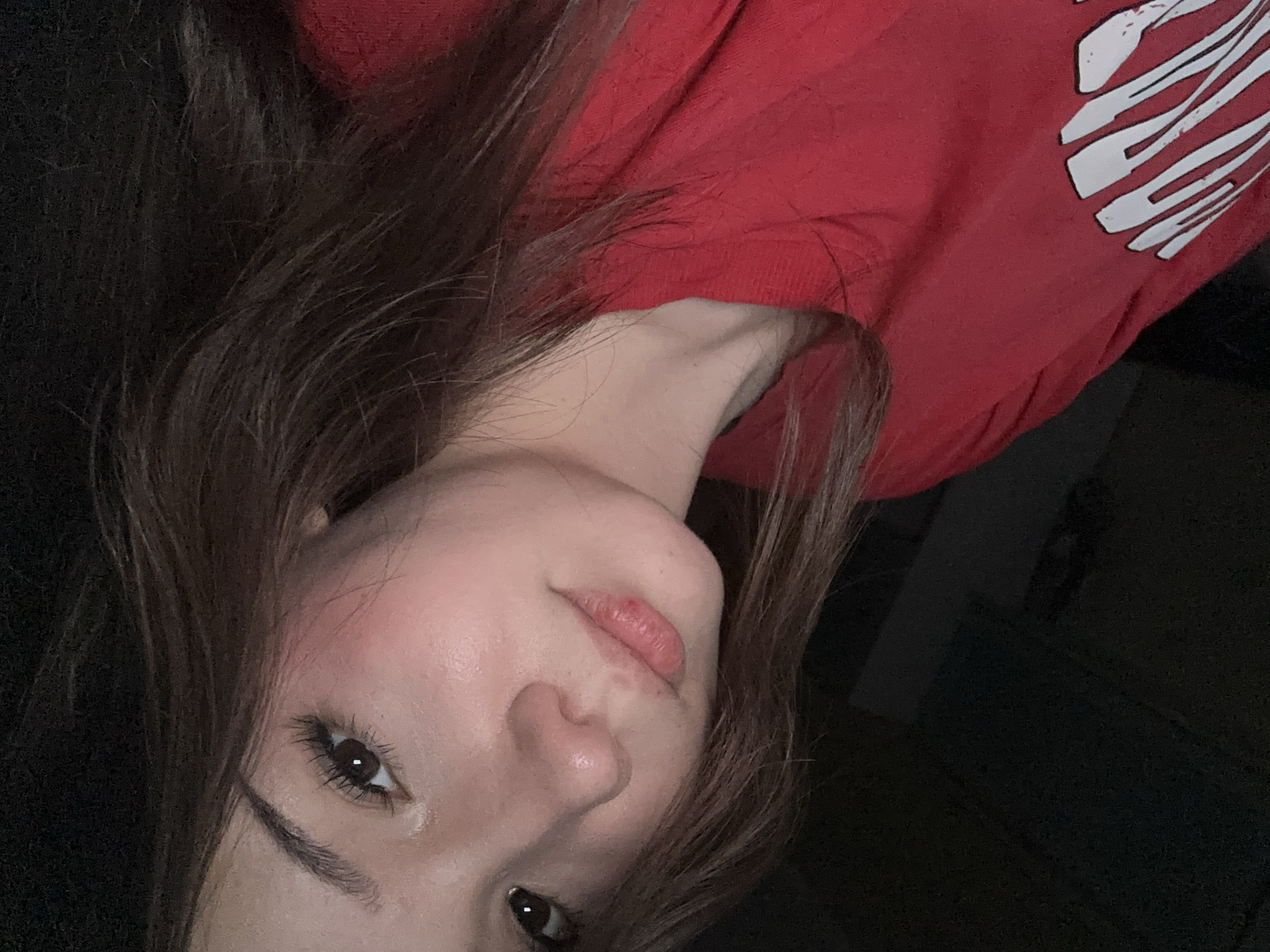 The image shows a close-up, head-and-shoulders portrait of a young woman with long, brown hair. She is wearing a red t-shirt with a white graphic design on the left side. Her skin is fair, with some blush on her cheeks. Her eyes are dark brown, and she has long eyelashes. Her lips are slightly parted and appear to have a small mark on the lower lip. The background is dark and out of focus, suggesting an indoor setting at night or in low light conditions. There are no visible landmarks or environmental cues to suggest a specific location within Naberezhnye Chelny, Russia. The overall impression is of a casual, informal selfie.