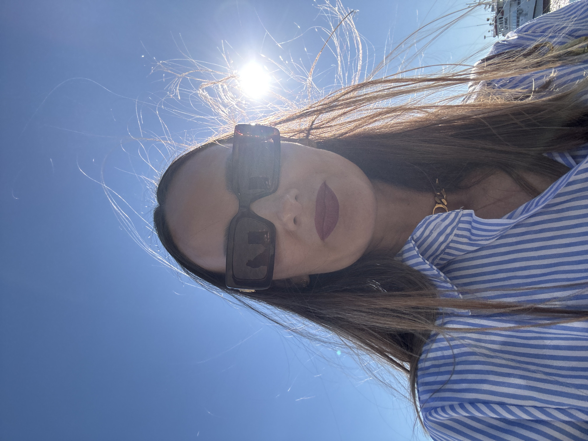 The media file shows a close-up, overhead shot of a woman against a bright blue sky. She is wearing large, dark sunglasses and a blue and white striped shirt. Her long, brown hair is being blown by the wind, with strands framing her face. Her lips are painted with a deep red lipstick. The sun is shining brightly, creating a lens flare effect and highlighting the windblown strands of her hair. In the background, a large white boat or ship is partially visible, suggesting the setting is near a body of water, likely a harbor. The overall impression is one of a sunny day, possibly at the coast, with a strong breeze. The upward angle of the shot and the bright sunlight create a dynamic and slightly abstract composition. No visible text is present in the image. The context provided suggests the location is Sochi, Russia, which is consistent with a seaside setting.