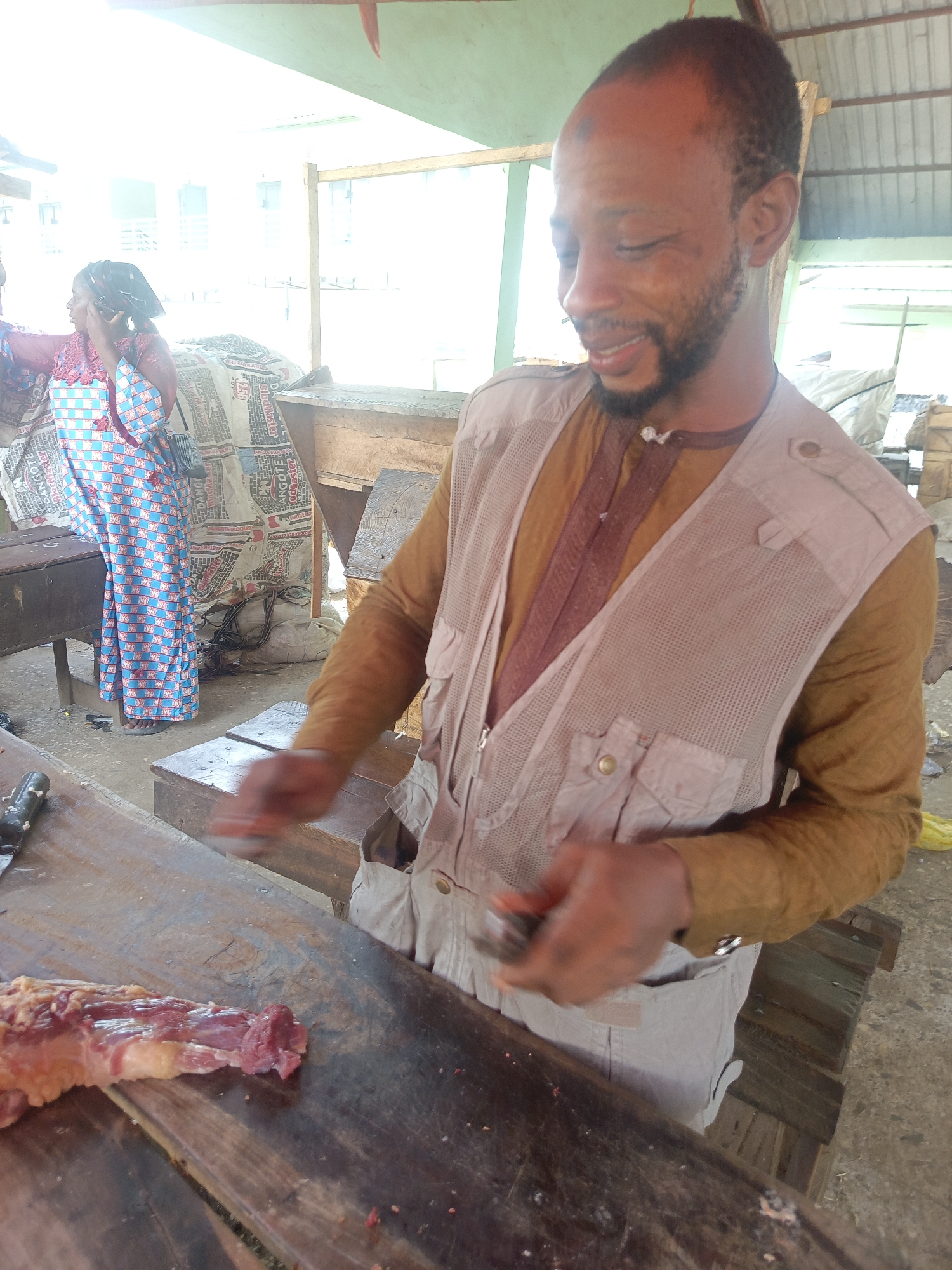 In Akwanga, Nigeria, a man stands behind a wooden table with a piece of raw meat on it. He is wearing a brown long-sleeved shirt and a light gray mesh vest. He is holding a small knife in his right hand and looking down at the meat with a slight smile. In the background, a woman in a blue and red patterned dress stands and talks on a cell phone. Behind her are various wooden structures and bags of cement. The scene is outdoors, possibly in a market or roadside butcher shop.