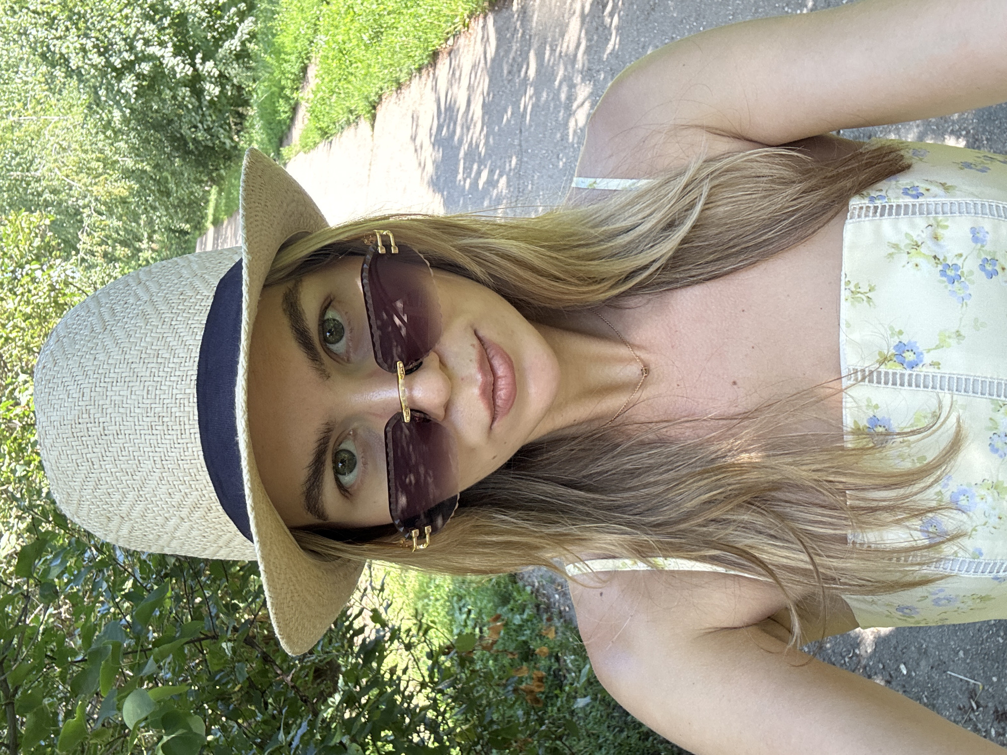 A woman wearing a straw hat, sunglasses, and a floral print dress is captured in a close-up selfie. She is outdoors, on a paved path lined with trees and grass. The path appears to be in a park or a wooded area within a city. The lighting suggests it is daytime. Her gaze is directed towards the camera, and she has a neutral expression.