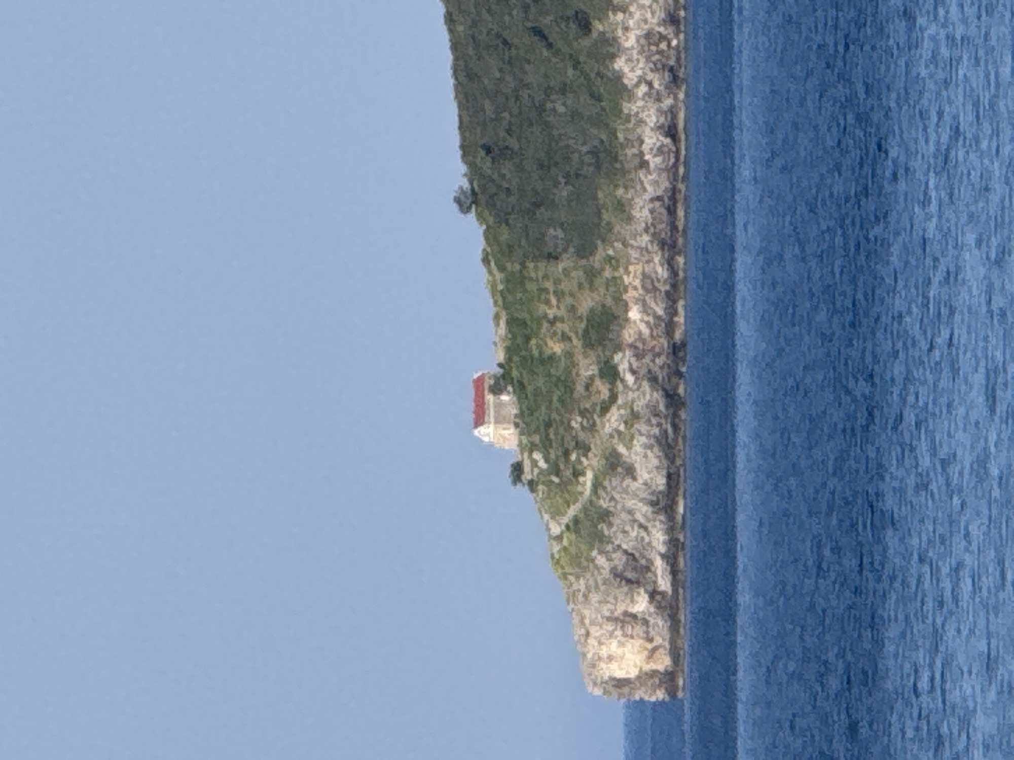 The image depicts a picturesque scene of a coastal cliff overlooking a body of blue water. The cliff is covered in lush green vegetation and rough, rocky terrain, suggesting a natural and somewhat wild landscape. Perched atop the cliff is a small, white building with a red roof, hinting at a dwelling or a historical structure. A path can be seen winding its way up the cliff towards the building, indicating access and a possible destination.
The sea is a deep blue, with a visible texture suggesting gentle waves or ripples. The sky is a clear, pale blue, indicating a bright day, likely with good weather. The orientation of the image is vertical, with the sky on the left and the sea on the right, and the cliff face running diagonally from the bottom left to the top right.
The overall impression is one of serenity and natural beauty, with the man-made structure adding a touch of human presence to the rugged landscape. There is no visible text in the image. The time of day appears to be daytime, given the bright lighting and clear sky. No specific location cues are evident, and the absence of people or any indication of activity suggests a peaceful and undisturbed setting.