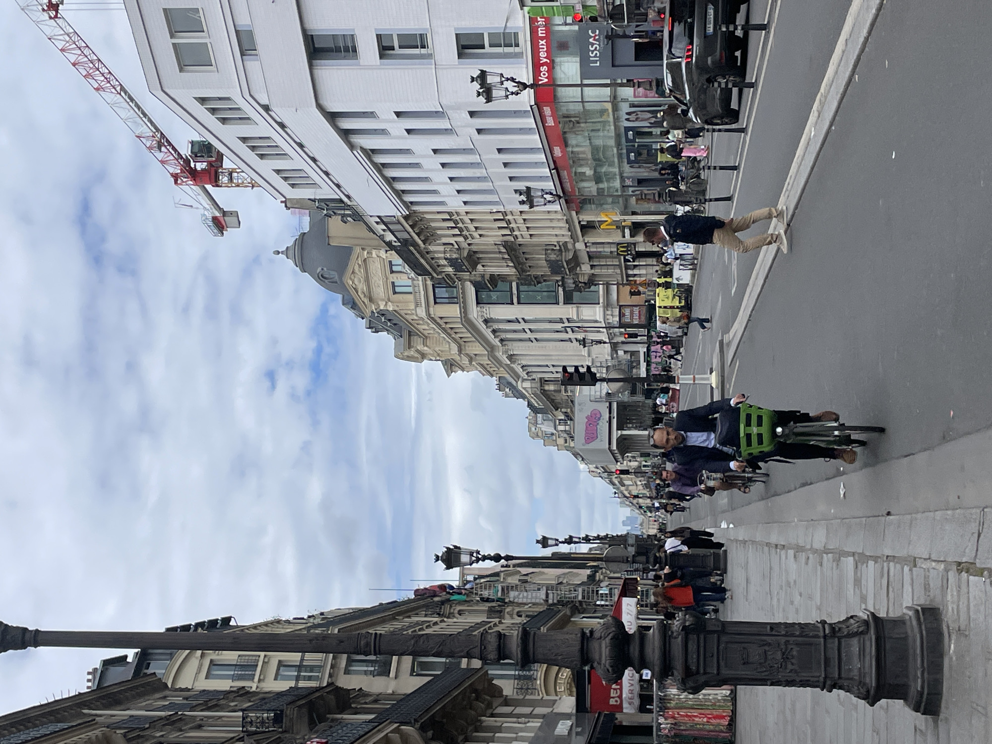 This is a street-level photograph taken in Paris, France, likely in a commercial or residential district. The image captures a busy street with pedestrians and cyclists, flanked by multi-story buildings with classic Parisian architecture. A prominent lamppost stands on the left side of the foreground, casting a shadow on the cobblestone sidewalk.
On the street, a man is riding a bicycle with a green basket, looking ahead. Behind him, other cyclists and pedestrians are visible. Several vehicles, including a dark-colored car, are present on the road. The buildings lining the street feature ornate facades, balconies, and large windows. A construction crane is visible in the background, suggesting ongoing development or renovation in the area. Traffic lights and signage are also present, indicating a functional urban environment. The sky is partly cloudy, suggesting overcast weather.