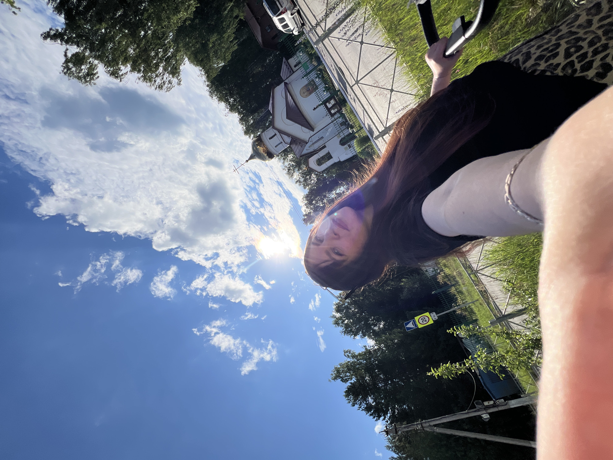 A woman with long, dark brown hair is taking a selfie outdoors in Noviy, Russia. She is wearing a black shirt and a leopard-print skirt. She is holding a stroller. In the background, there is a white church with a golden dome, surrounded by trees. The sky is blue with scattered clouds, and the sun is shining. A speed limit sign with the number 20 is visible on the left side of the image. The overall scene suggests a sunny day in a semi-rural or suburban area.