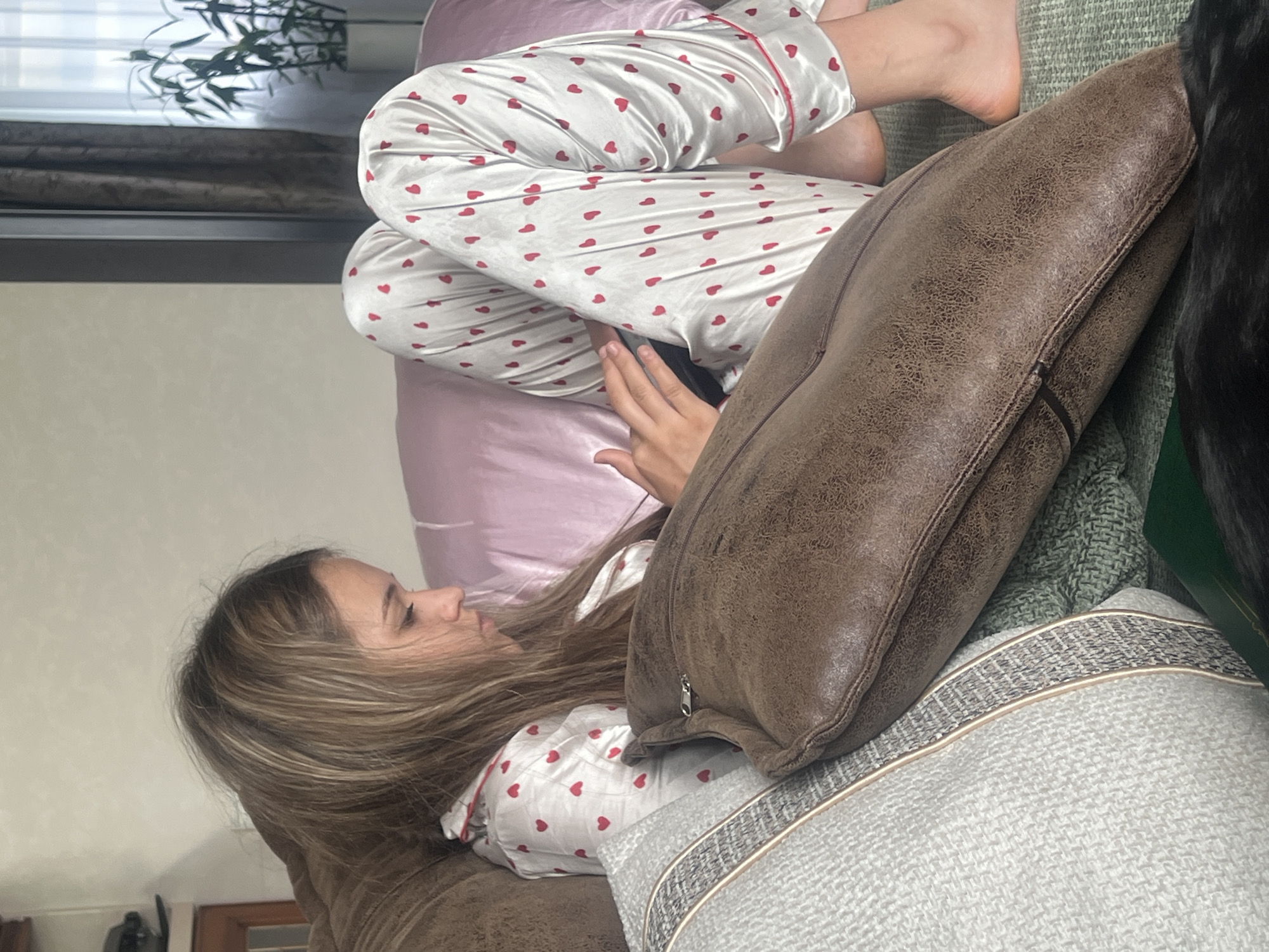 A young woman with long, blonde hair is lying on a couch, looking down at her phone. She is wearing white pajamas with red hearts. The couch is covered in pillows and blankets. In the background, there is a window with curtains and a plant. The lighting is soft and natural. The scene appears to be taking place in a home.