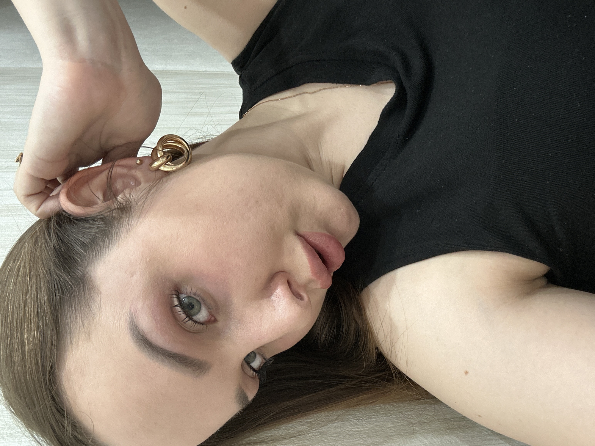 A young, fair-skinned woman lies on a light-colored surface, likely the floor. She wears a black top and a gold-colored earring with a multi-loop design. The woman has light brown hair and green eyes. Her lips are slightly parted. One of her hands is near her ear. There's a gold necklace around her neck. There is no information available to infer that the location is in Chaikovsky, Russia.