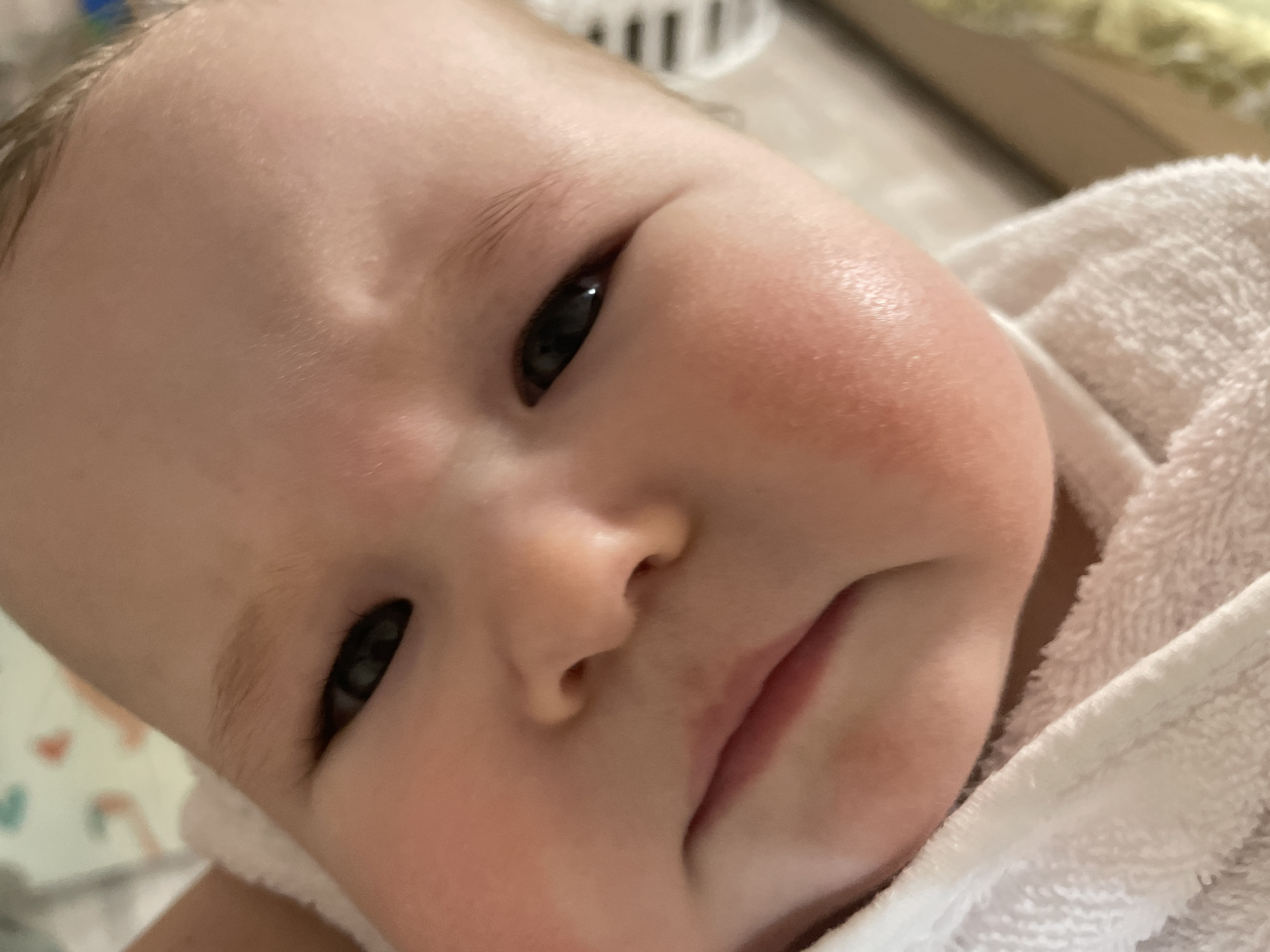 A close-up photograph shows a baby's face. The baby has fair skin, dark eyes, and slight redness on the cheeks. The baby's expression appears serious, with a furrowed brow and pursed lips. A white towel is draped around the baby. The location is unknown and there is no indication it is in Chaikovsky, Russia.