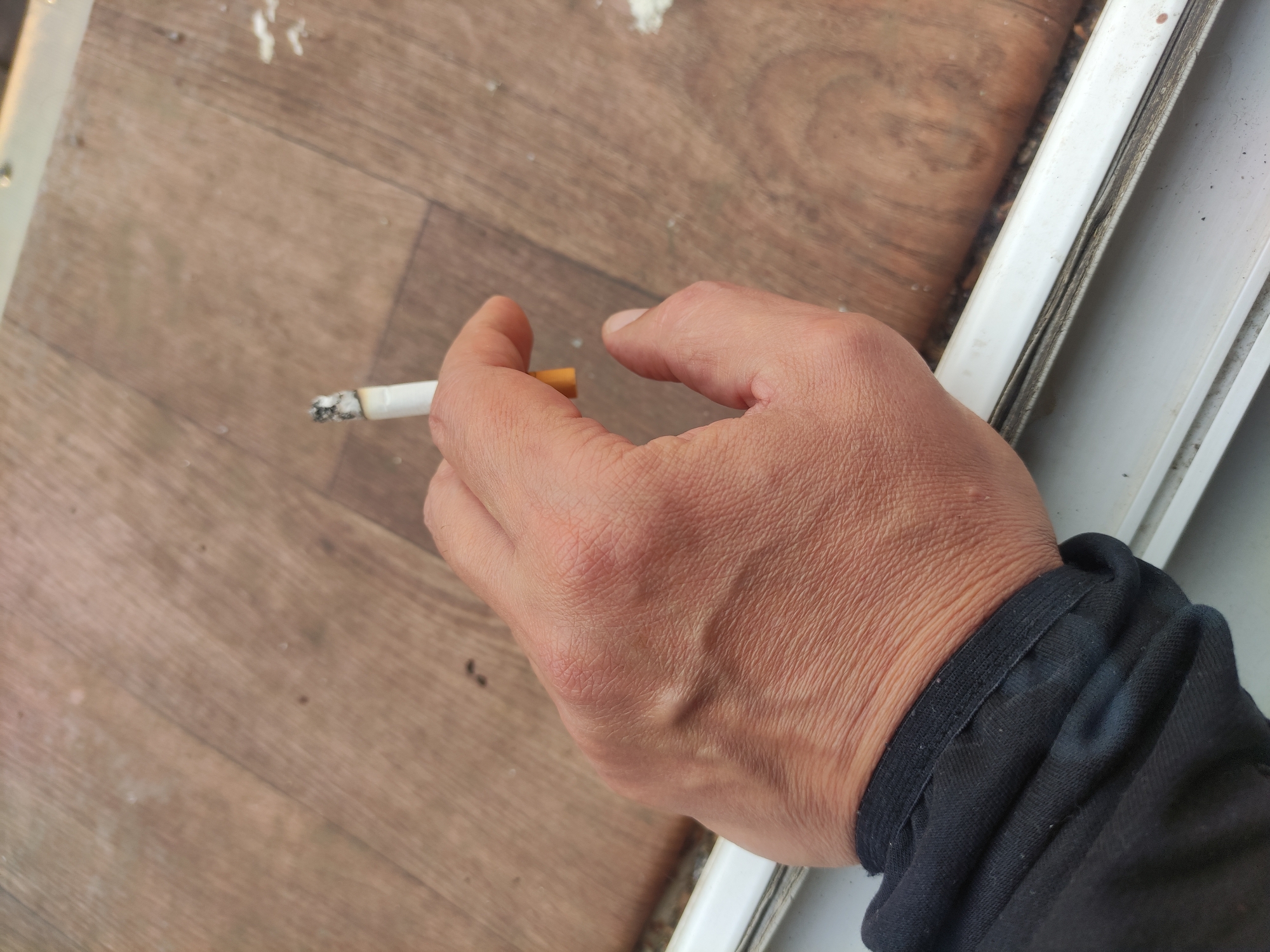 A lit cigarette is held between the fingers of a light-skinned hand with prominent veins. The hand is wearing a black cuff. The background features faux-wood flooring and part of a white frame. The location and city cannot be determined from the image.