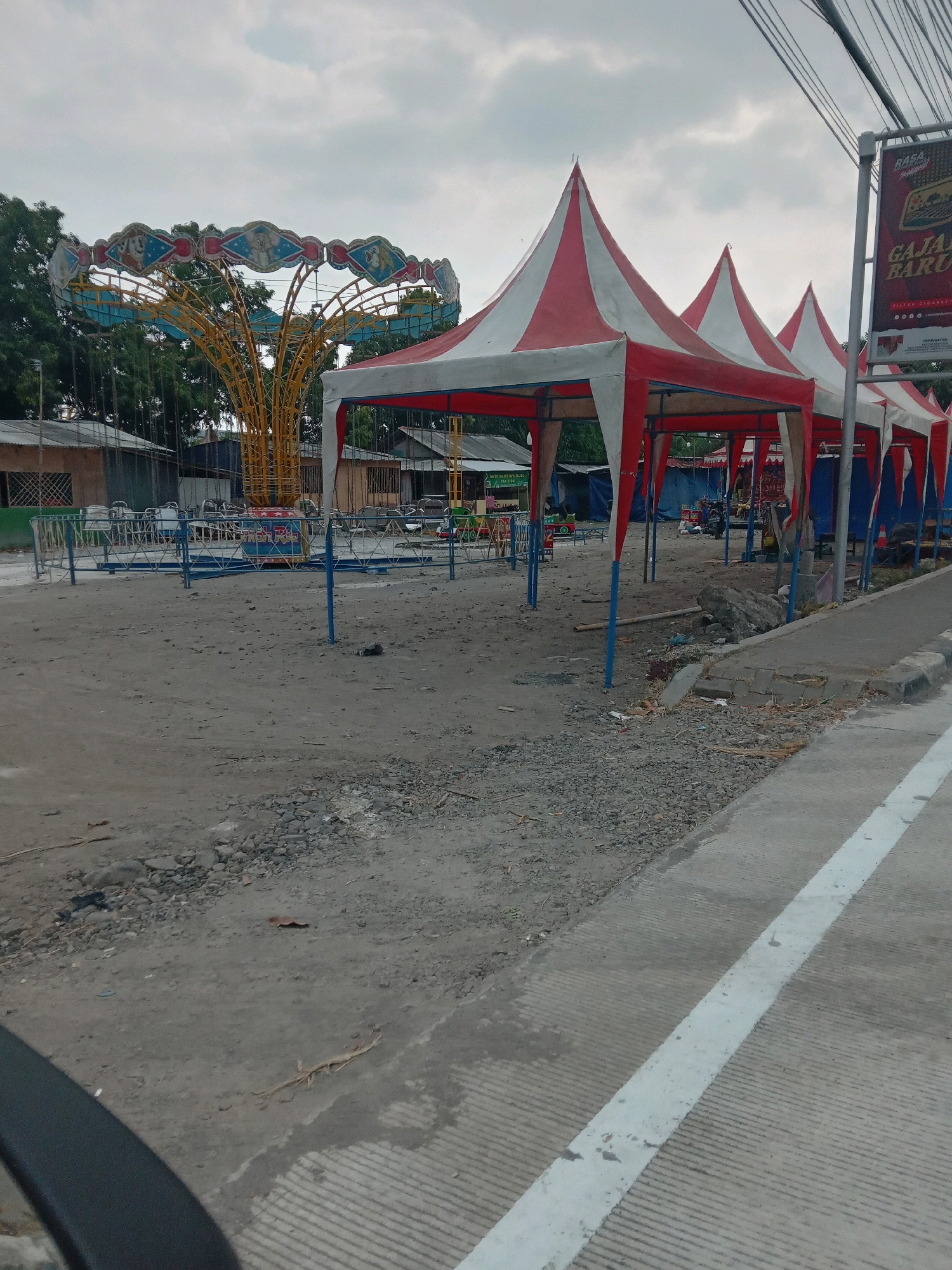 The image depicts an outdoor scene in Srimulyo, Indonesia. In the foreground, a paved road with a white dividing line runs diagonally from the bottom right to the top left. To the left of the road is a dirt and gravel area. Several red and white striped tents with conical roofs are set up in the middle ground, supported by blue poles. Behind the tents, an amusement park ride with a yellow central tower and a circular seating area adorned with images of animals is visible. In the background, there are simple buildings, trees, and electrical wires overhead. A sign with text in Indonesian is positioned to the right of the tents. The sky is overcast with clouds. No people are visible in the image. The scene appears to be an amusement area that is either temporarily set up or not currently in operation.