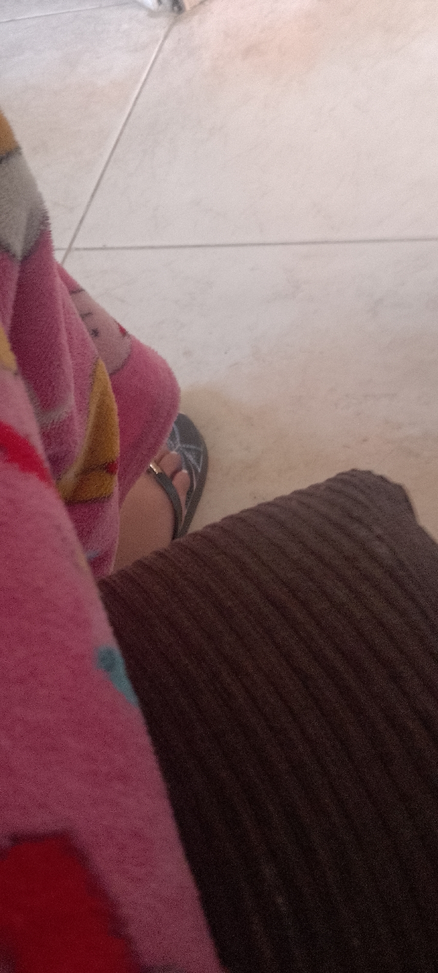 The image shows a close-up view of a person's legs and feet, resting on what appears to be a dark brown, corduroy-like surface. The person is wearing a pink, fuzzy blanket with a pattern that includes cartoon characters and yellow accents. On the feet, there are dark gray flip-flops with a gold accent on the strap. The background is a tiled floor, with light-colored tiles and visible grout lines. The lighting suggests it might be daytime. The scene is indoors, likely in a home, given the cozy blanket and casual footwear. There is no visible text in the image. The overall impression is one of relaxation and comfort.