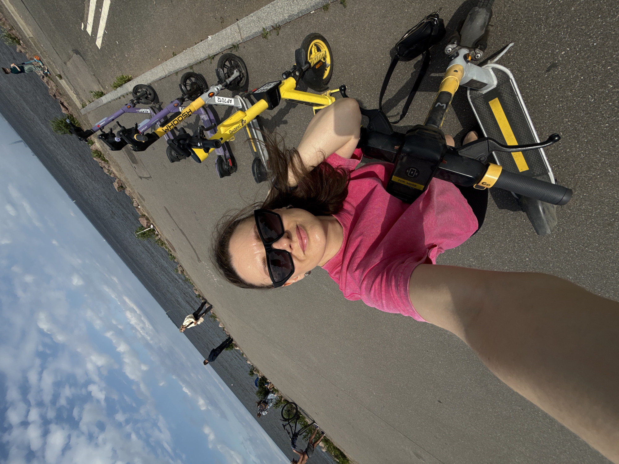 A woman in a pink shirt and sunglasses is taking a selfie while standing next to a row of yellow and purple electric scooters. The scooters are parked on a paved walkway next to a large body of water, likely the Gulf of Finland. In the background, several people are walking and one person is standing near a bicycle. The sky is blue with scattered clouds. The scene appears to be located along a waterfront promenade in Saint Petersburg, Russia.