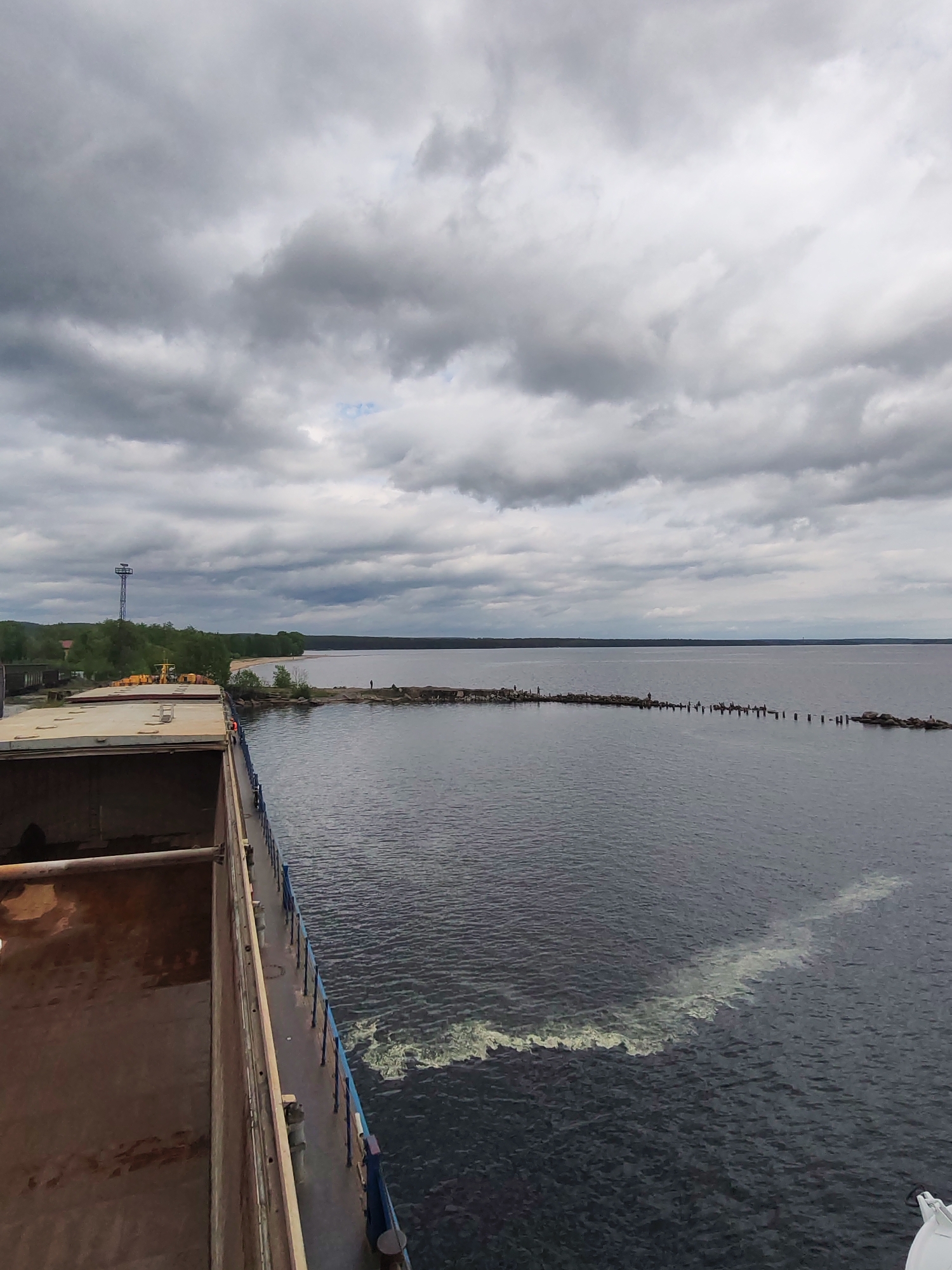 This wide-angle photograph captures a vast maritime or lakeside scene in Medvezhyegorsk, Russia, viewed from the deck of a large vessel, likely a cargo ship or barge. The ship's empty, rust-colored cargo hold and a section of its blue railing are visible in the left and bottom foreground. A white, foamy wake trails behind the moving vessel in the dark, rippled water.
The middle ground is dominated by a large body of calm, dark grey water, presumably Lake Onega, extending towards a distant, tree-lined coastline. A long, low breakwater constructed from rocks and interspersed with wooden posts stretches across the water, originating from the land and curving gently to the right. Several small human figures are discernible on this breakwater, suggesting people are fishing or observing the water.
On the shoreline to the left, green trees partially obscure industrial or port infrastructure, including yellow machinery and dark buildings. A tall, blue metal tower, possibly a navigation aid or communication mast, stands prominently above the trees. Further along the coast, a sandy or rocky beach is visible, bordered by a dense line of trees.
The sky is heavily overcast with thick, uniform grey clouds, indicating a cloudy or gloomy daytime. No direct sunlight is visible, contributing to a muted color palette. The scene conveys a sense of quiet vastness at the edge of a large body of water. No discernible text is present in the image.
