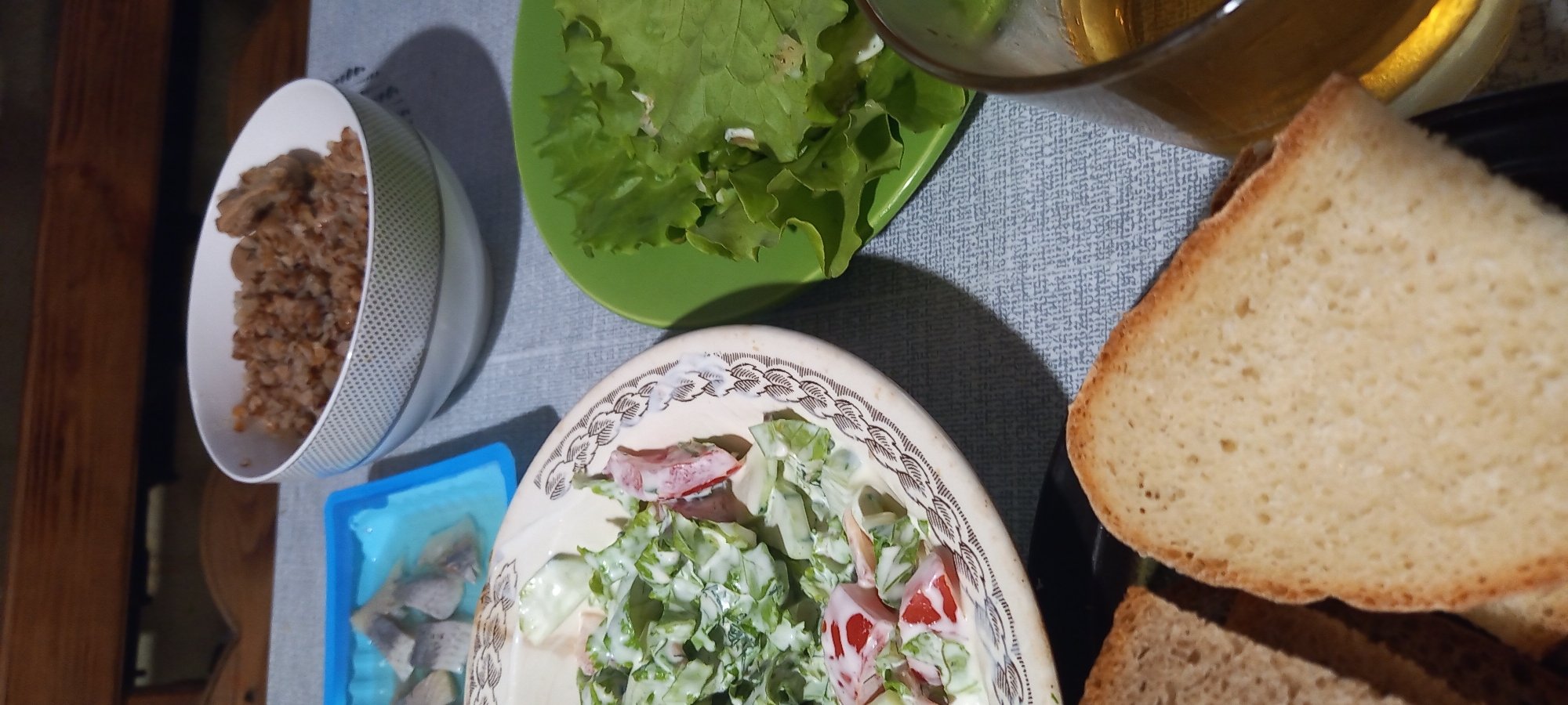 A close-up, high-angle shot captures a meal laid out on a grey, textured tablecloth. In the background, a wooden surface suggests a table or a bench. The foreground is dominated by slices of bread. To the left, two slices of toasted bread are visible, with one partially obscured. To the right, a larger slice of bread is prominently featured, its texture revealing small air pockets.
Moving towards the middle ground, a creamy salad is presented in a beige bowl with an intricate, dark brown patterned rim. The salad appears to contain chopped lettuce, cucumber, and tomatoes, all coated in a white dressing. Next to the salad, a vibrant green plate holds fresh lettuce leaves.
In the upper left quadrant, a blue plastic container holds pieces of pickled herring. Behind this, a white bowl filled with buckwheat and what appears to be pieces of meat or mushrooms sits on the tablecloth. To the far right, a glass filled with a golden liquid, likely a beverage, is partially visible.
The scene appears to be an indoor dining setting, possibly a home, and the presence of various food items suggests a meal in progress. The lighting is soft and diffused, casting subtle shadows. The location of this scene is not specified within the image itself, and therefore, its presence in Kryvyi Rih, Ukraine cannot be determined from the visual information provided.