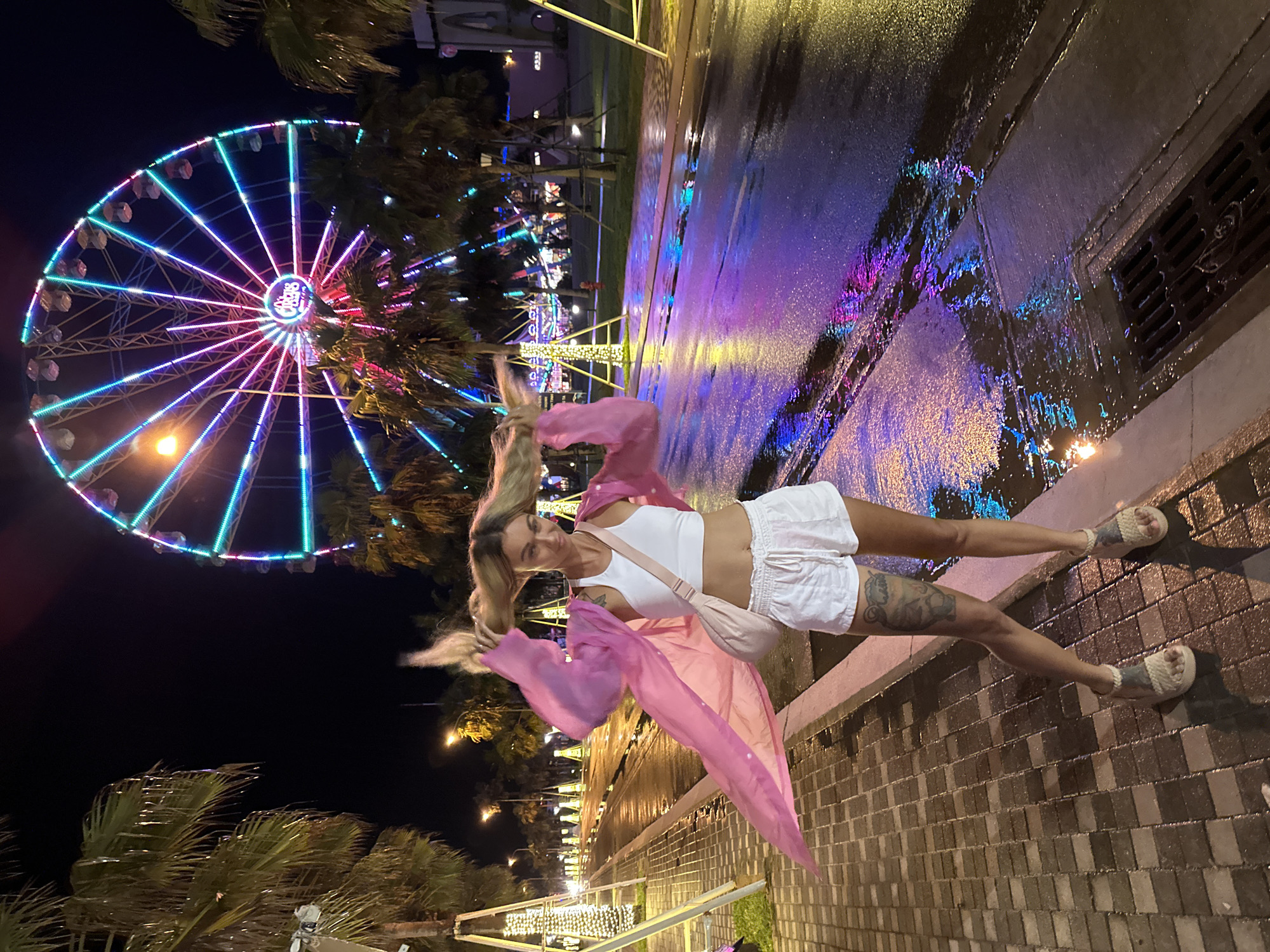This night-time photograph, set in Tien Thanh Ward, Vietnam, features a woman posing playfully on a wet street next to a brightly lit Ferris wheel. The woman, with long blonde hair in motion, wears a white crop top, white shorts, and a flowing sheer pink cover-up. She sports light-colored sandals and has tattoos visible on her legs. She is smiling and looking towards the camera, conveying a cheerful mood.
The prominent background element is a large Ferris wheel, illuminated with vibrant blue, pink, and purple lights, whose central hub clearly displays the word "Circus." Palm trees are visible throughout the scene, interspersed with other decorative lights and streetlamps, indicating an entertainment or promenade area. The ground, a mix of asphalt and brick paving, is visibly wet, creating vivid reflections of the colorful lights from the Ferris wheel and other distant attractions. The overall setting is brightly lit by artificial lights under a dark night sky, suggesting a lively evening atmosphere.