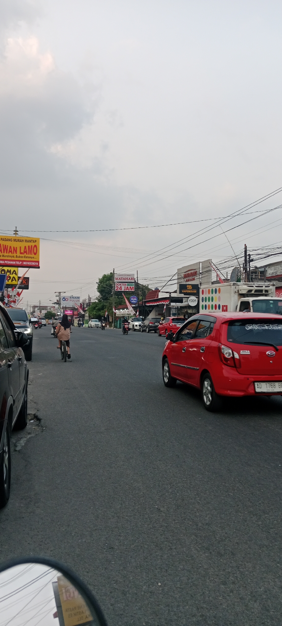 The image captures a street scene in Maguwoharjo, Indonesia, under an overcast sky. A two-lane asphalt road runs through the center of the frame, with vehicles in motion and parked on either side. On the left side of the road, a black SUV is partially visible, followed by a red car. Further down the road, a person is riding a bicycle. On the right side of the road, a red hatchback is prominent, its license plate reading "AD 1768 ER". Behind the red car, several other vehicles, including a white truck, are visible.
Billboards and signs are a significant feature of the environment. On the left, a large yellow billboard reads "PADANG MURAH MANTAP LAWAN LAMO". Other signs with Indonesian text are also present. On the right, a sign for "MATAHARI DOKTER 24 JAM" is visible, as is a sign for "VAPEHOUSE". The buildings along the roadside appear to be commercial establishments. Electrical wires crisscross overhead. The overall impression is a busy, commercial area in an Indonesian city.