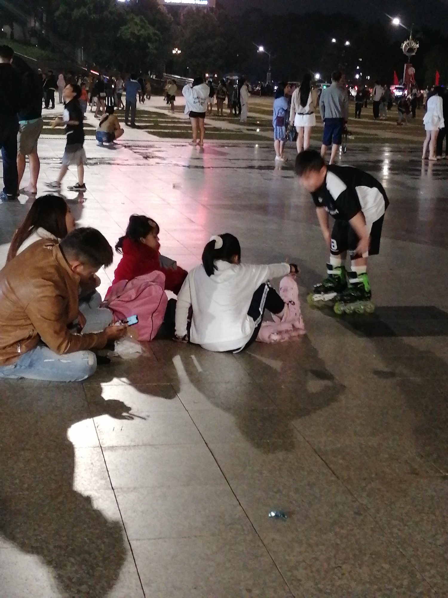 The image captures an outdoor scene at night in what appears to be a public square or promenade in Da Lat, Vietnam, indicated by the style of the paving stones and the ambient lighting which suggests a tourist area. The ground is wet and reflective, possibly from recent rain, creating distorted shadows from the people present.
In the foreground, a group of individuals are seated on the ground. On the left, a man wearing a brown leather jacket and blue jeans is looking down at a smartphone he is holding. Beside him, a woman with long dark hair is also seated. To their right, a young child in a red top is sitting with a pink bag. Further to the right, a child in a white hooded jacket is seated, facing away from the camera, and a pink blanket or piece of fabric is near her.
In the mid-ground and background, there are many other people scattered throughout the area, some walking and others standing. One young boy is wearing rollerblades and is in a crouched position, interacting with the child in the white jacket. He is wearing a black and white shirt and black shorts. The overall atmosphere is one of a bustling evening gathering in a city. The lighting suggests artificial illumination from streetlights and possibly lights from nearby buildings, creating a slightly hazy effect.