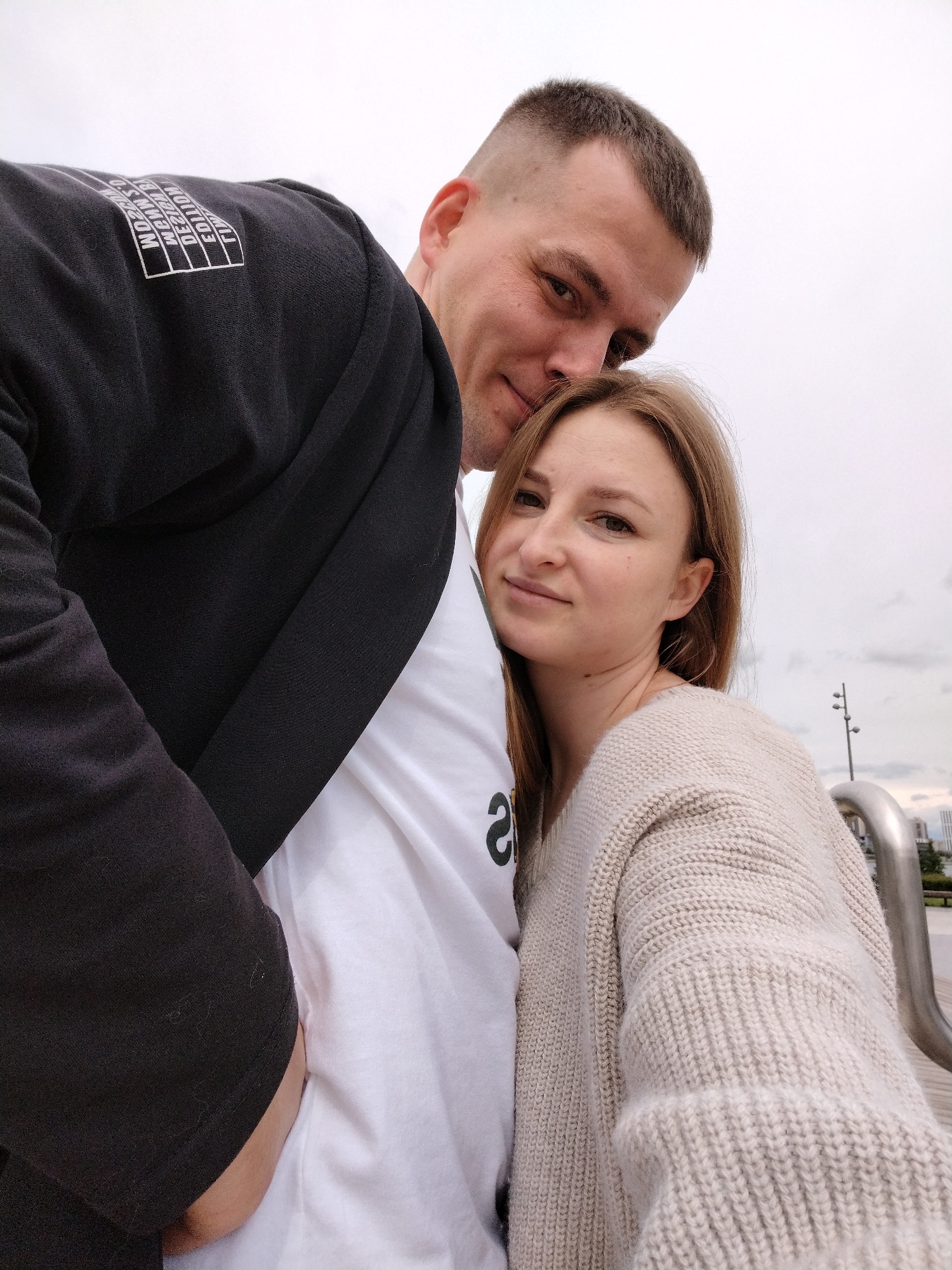 Two individuals, a man and a woman, are captured in a photograph taken outdoors. The man, on the left, has short hair and is wearing a black jacket over a white t-shirt. A rectangular patch with text is visible on the back of his jacket. He is leaning his head towards the woman. The woman, on the right, has shoulder-length light brown hair and is wearing a light-colored sweater. They are standing close to each other. In the background, there are metallic structures resembling railings and distant buildings under a cloudy sky. Based on available information, the location is inferred to be in Kazan, Russia.