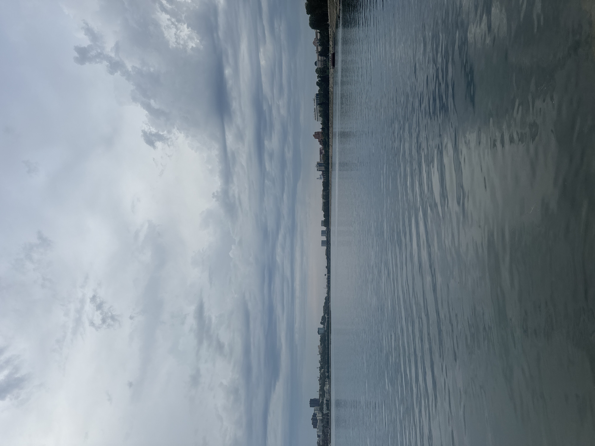 The image shows a wide, calm body of water, likely a river or lake, reflecting the overcast sky. On the far bank, a cityscape is visible with a mix of modern high-rise buildings and older, lower structures. Trees line the waterfront in some sections. The sky is filled with heavy, gray clouds. This scene is consistent with the waterfront of the Angara River in Irkutsk, Russia, where the city's skyline meets the water. There are no people or specific actions depicted. The overall mood is tranquil and somewhat somber due to the cloudy weather.