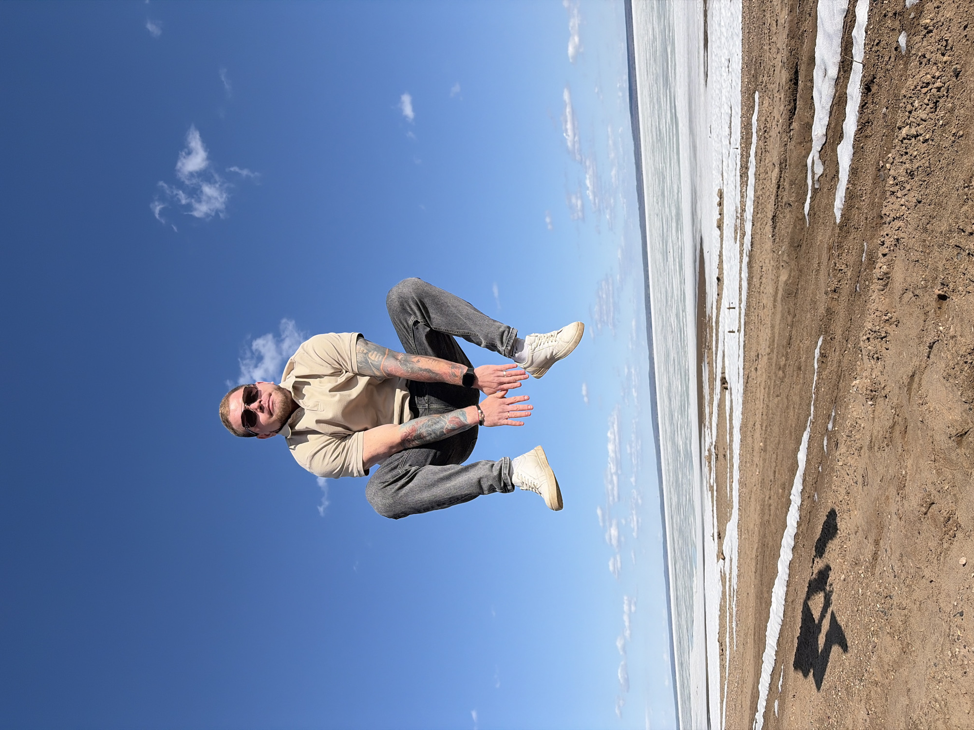 A man wearing a beige shirt, gray jeans, white shoes, and sunglasses is in mid-air against a blue sky with some clouds. He has tattoos on his arms. The landscape is on the right side of the photo and consists of a frozen river. The location is the city of Noviy, Russia.