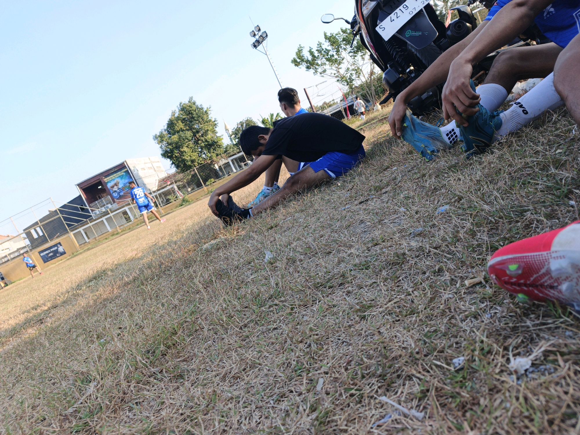 The image shows a group of people on a dry, grassy field, likely a sports field, in Mojokerto, Indonesia. In the foreground, a person is sitting and stretching their legs, wearing a black t-shirt, blue shorts, white socks, and blue soccer cleats. To the right, another person's legs are visible, also wearing white socks and one person's leg is wearing a red soccer cleat. In the background, two individuals are standing, one wearing a blue jersey with the number 26. A motorcycle with a license plate is parked to the right of the frame. A building with a mural is visible in the distance, along with a soccer goal and floodlights. The sky is clear and blue, indicating a sunny day.