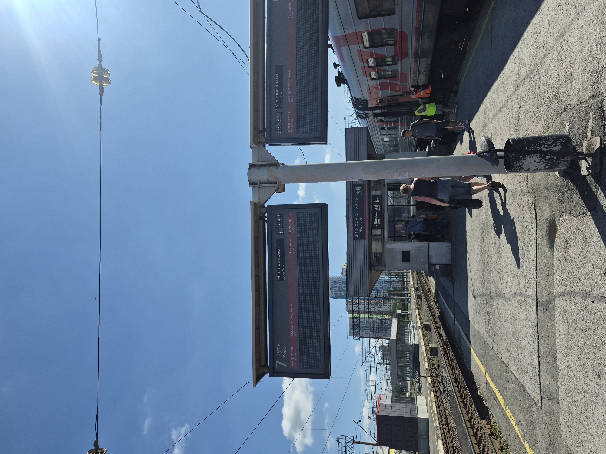 The image depicts a sunny day at a train station platform in Yekaterinburg, Russia. In the foreground, a train with a red and gray livery is stopped at the platform. Several individuals are visible on the platform, including passengers walking and station personnel. Two digital information displays are mounted on a pole, providing train schedule information, with one showing "Track 7" and the other displaying "Local time 18:42". The environment includes railway tracks, overhead power lines, modern station buildings in the background, and a clear blue sky with scattered clouds. The platform surface is a mix of concrete and asphalt with a yellow line marking the edge. A trash receptacle is also visible near the pole.