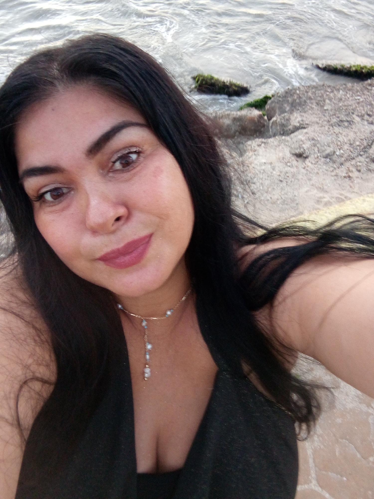 A selfie of a dark-haired woman with light-brown eyes wearing a gold necklace with a light-colored pendant. She is standing at a rocky beach in Juan Griego, Venezuela, with seawater in the background.
