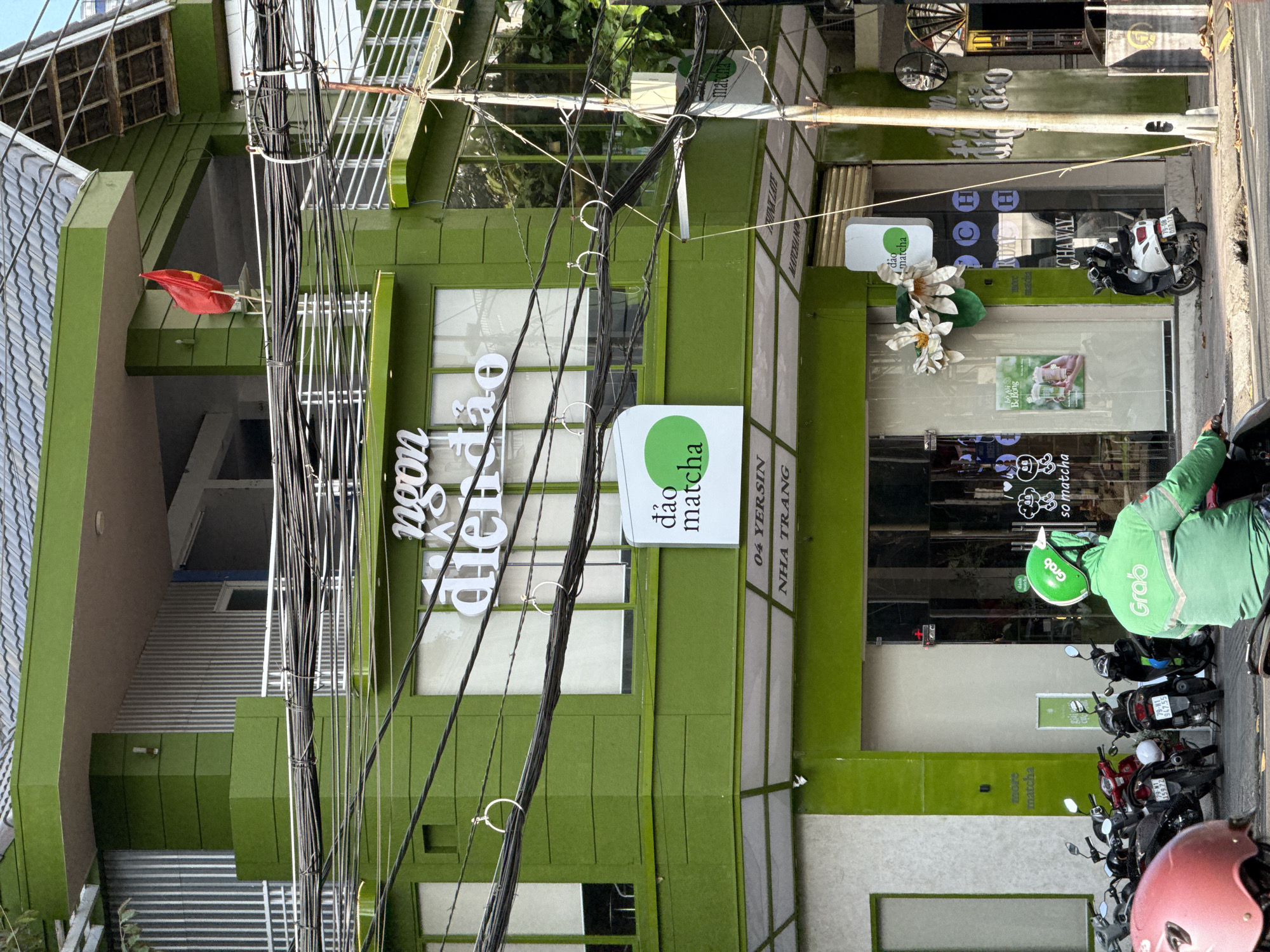 The image depicts the exterior of "Dao Matcha," a matcha shop located at 04 Yersin Street in Nha Trang, Vietnam. The building is painted green and features the shop's signage in Vietnamese and English. Several motorcycles are parked in front, with one Grab delivery driver visible wearing a green helmet and jacket, waiting on his scooter. The shop's window displays promotional material and decorative flower arrangements. A Vietnamese flag is visible on the upper level of the building. Overhead, a dense network of power lines crisscrosses the sky.