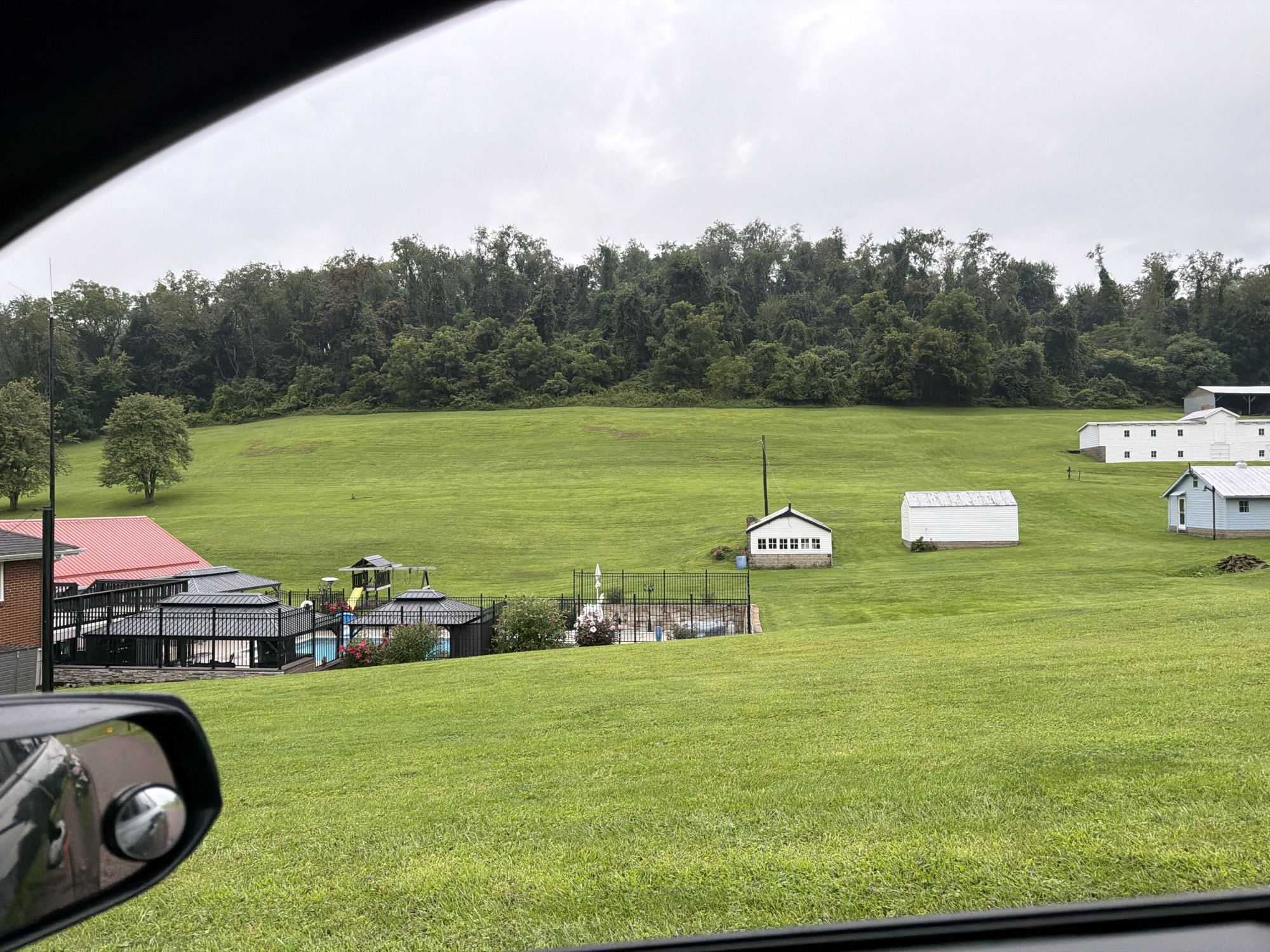 The image shows a rural landscape with several buildings and recreational facilities on a cloudy day. In the foreground, a car's side mirror is visible on the left, reflecting a portion of the car's interior. The main scene depicts a large, gently sloping green field with a dense line of trees forming the horizon.
Several structures are scattered across the field. On the left side, a building with a red roof is partially visible, connected to a fenced area with multiple covered structures, likely gazebos, and an in-ground swimming pool. A playground set with a slide is also present within this fenced area.
Further into the field, there are several smaller buildings. A white building with a dark roof and visible windows is situated on a slight rise. To its right and slightly further away, another long, white building with a weathered roof is visible. To the far right, a light blue building with a white roof and dark-framed windows is present. In the distance, a larger white building with a white roof and multiple dark-framed windows is also seen.
The environment appears to be a well-maintained property with ample open green space. The sky is overcast, suggesting a dull or possibly rainy weather condition. The overall impression is that of a residential or recreational property in a rural setting. The image appears to be taken from within a vehicle, looking out. This scene is located in Fallowfield Township, United States of America.