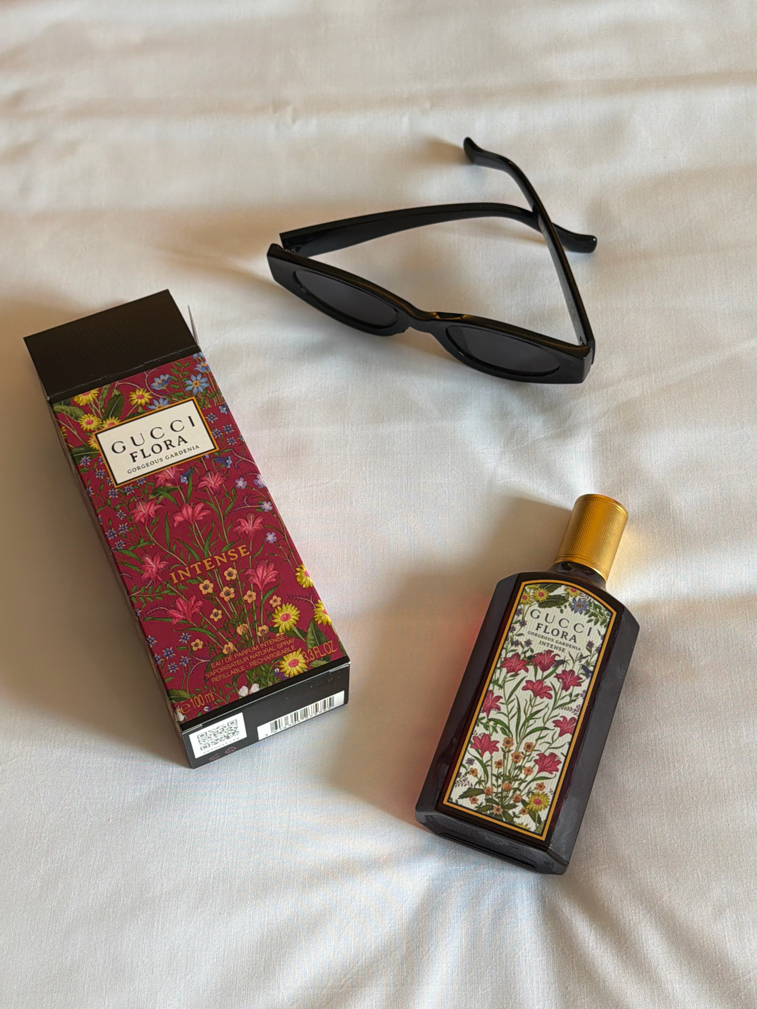 The image shows a flat lay of a Gucci Flora Gorgeous Gardenia Intense perfume bottle and its box, along with a pair of black sunglasses, all arranged on a white, textured surface, likely a bedsheet. The perfume box is positioned to the left, showcasing a floral pattern with pink, yellow, and blue flowers against a deep red background, and text including "GUCCI FLORA" and "INTENSE". To the right of the box is the perfume bottle, also featuring the same floral design on its label and a gold cap. The black sunglasses are placed in the upper right quadrant of the image, with their arms crossed. The overall lighting suggests an indoor setting, possibly a bedroom, with soft, diffused light. The scene does not contain any elements that indicate its location in Breda, Netherlands, or any specific urban environment.