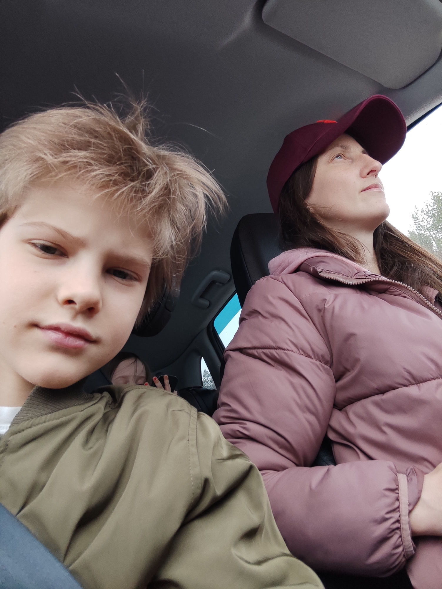 The photograph shows three individuals inside a vehicle. A young boy with blond hair is in the foreground, wearing an olive green jacket. To his right, a woman is wearing a burgundy baseball cap and a pink puffer jacket, looking upwards. Partially visible in the background between the two is another person. The interior of the vehicle is visible. The location is possibly in Maksimovo, Russia.