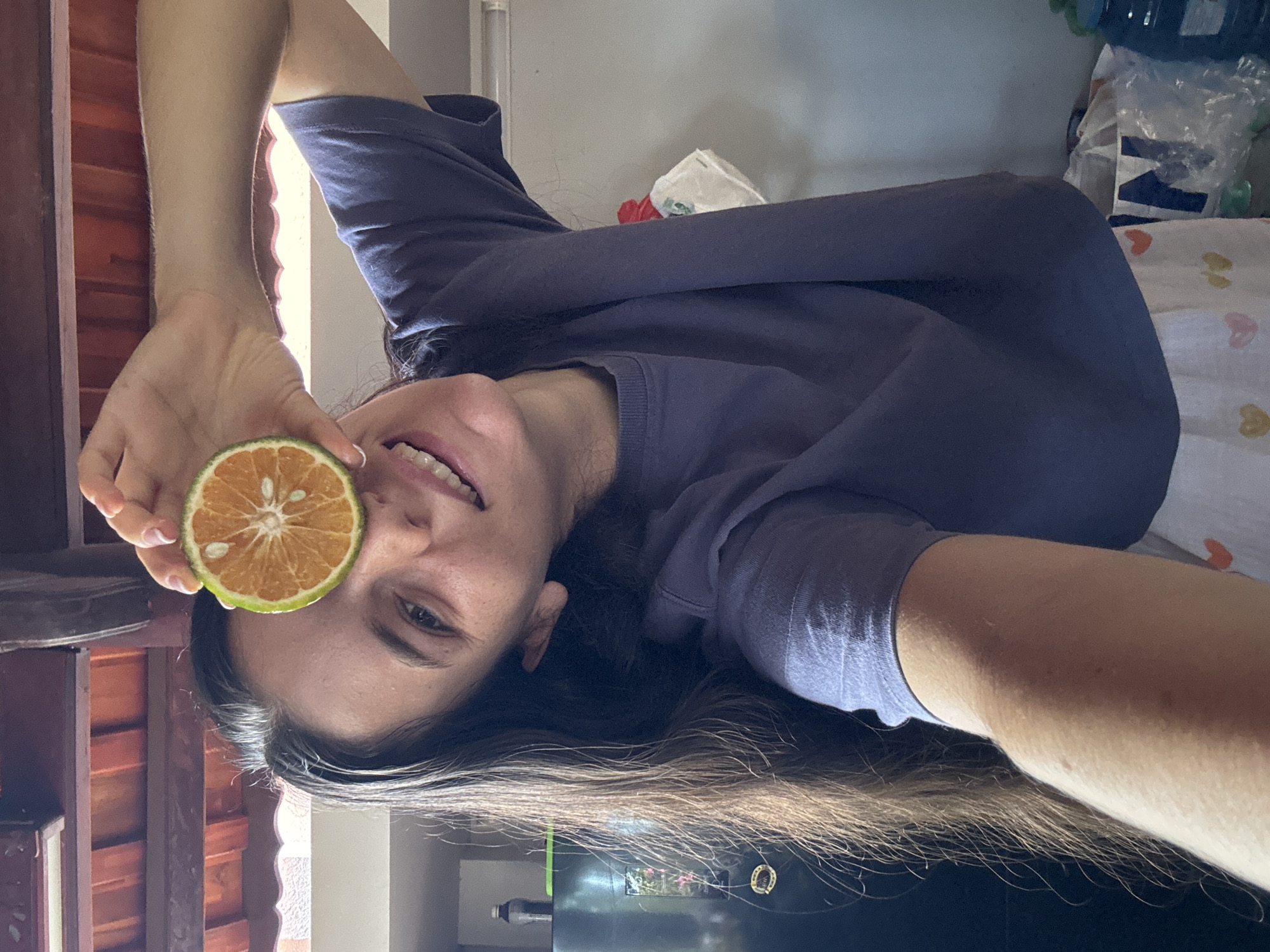 A woman holds a halved orange up to her eye. She is smiling. She is wearing a purple t-shirt and is lying down. Her long brown hair spreads out beneath her. Behind her are some items on a table, including a plastic bag. The location is Mũi Né, Vietnam.