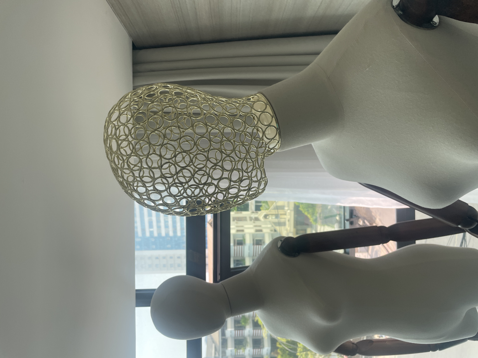 The image shows two mannequins. The first mannequin wears a gold, spherical wire headpiece. Both mannequins are off-white, with dark wooden arms and shoulders. The scene is indoors, near a window offering a view of Bắc Nha Trang, Vietnam, characterized by tall buildings and greenery.
