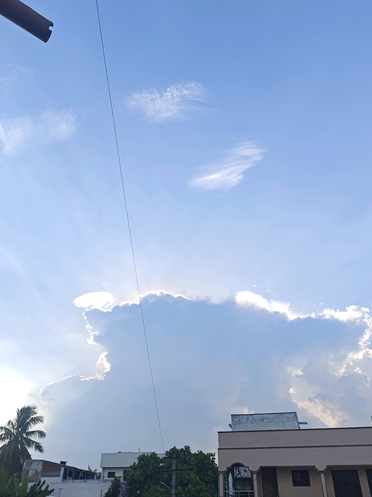 The image depicts a bright sky with scattered clouds, taken in Nellore, India. A dark, thin wire stretches diagonally across the frame from the top left corner, disappearing into the sky. In the lower portion of the image, buildings and greenery are visible. A palm tree is on the left, and a modern-looking building with a cream-colored facade and brown trim is on the right. The sky is mostly blue with significant cloud cover, particularly a large, bright cloud formation illuminated by sunlight, creating a halo effect. The perspective suggests the photo was taken from an elevated position, possibly a rooftop.