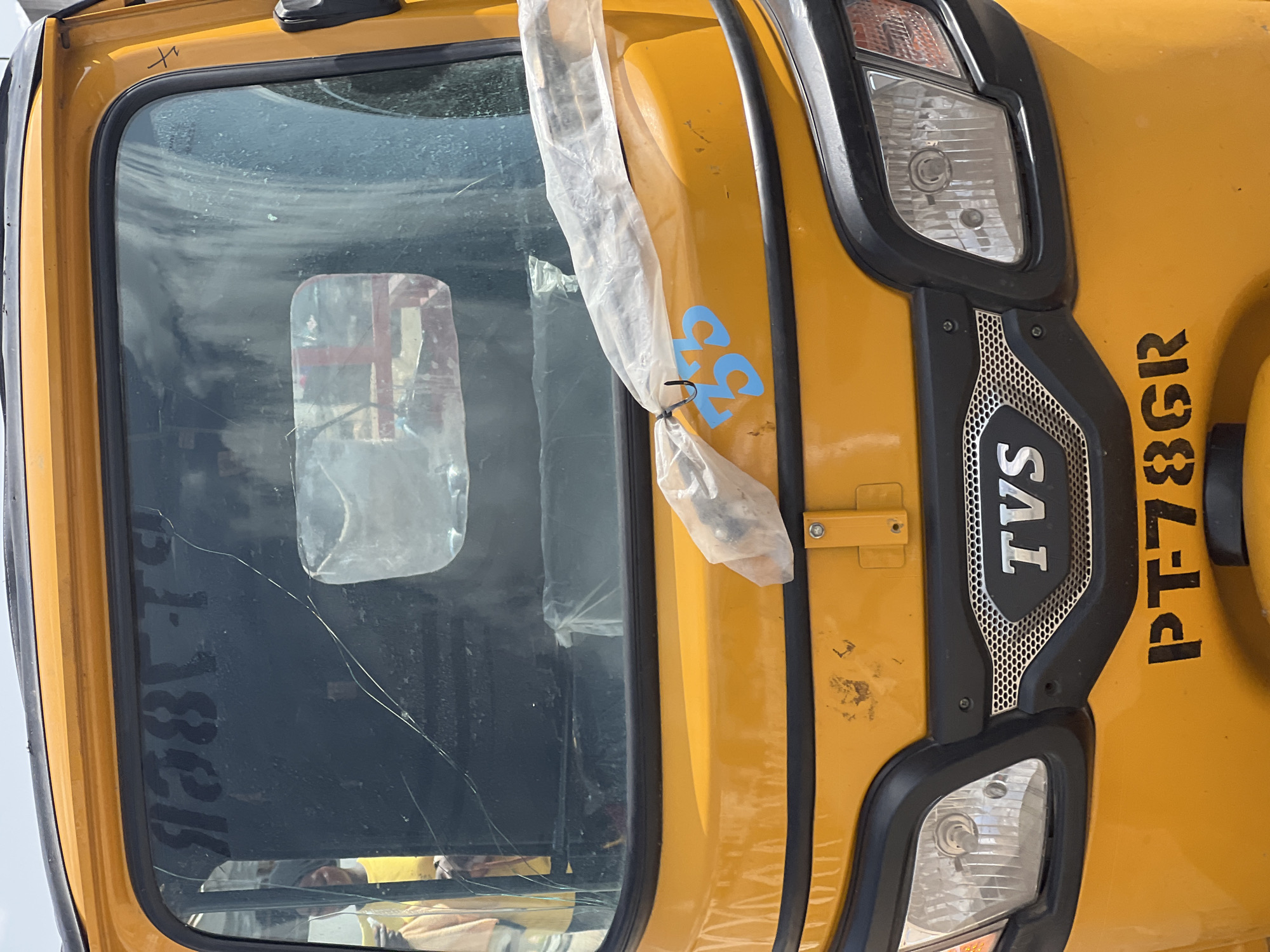 This is a close-up, eye-level photograph of the front of a yellow auto-rickshaw, commonly known as a "keke." The vehicle's windshield is cracked and reflects the sky and surrounding structures. A piece of clear plastic is taped to the windshield, obscuring part of the view. A blue number "35" is visible on the yellow bodywork below the windshield. The headlights and a black grille with the "TVS" logo are prominent. The license plate, "PT-78GR," is stenciled in black on the lower front of the vehicle. The scene appears to be an outdoor setting, likely a street or parking area, given the natural lighting and the context of the vehicle. The image does not provide enough information to definitively place it in Jalingo, Nigeria, but the type of vehicle is common in such regions.
