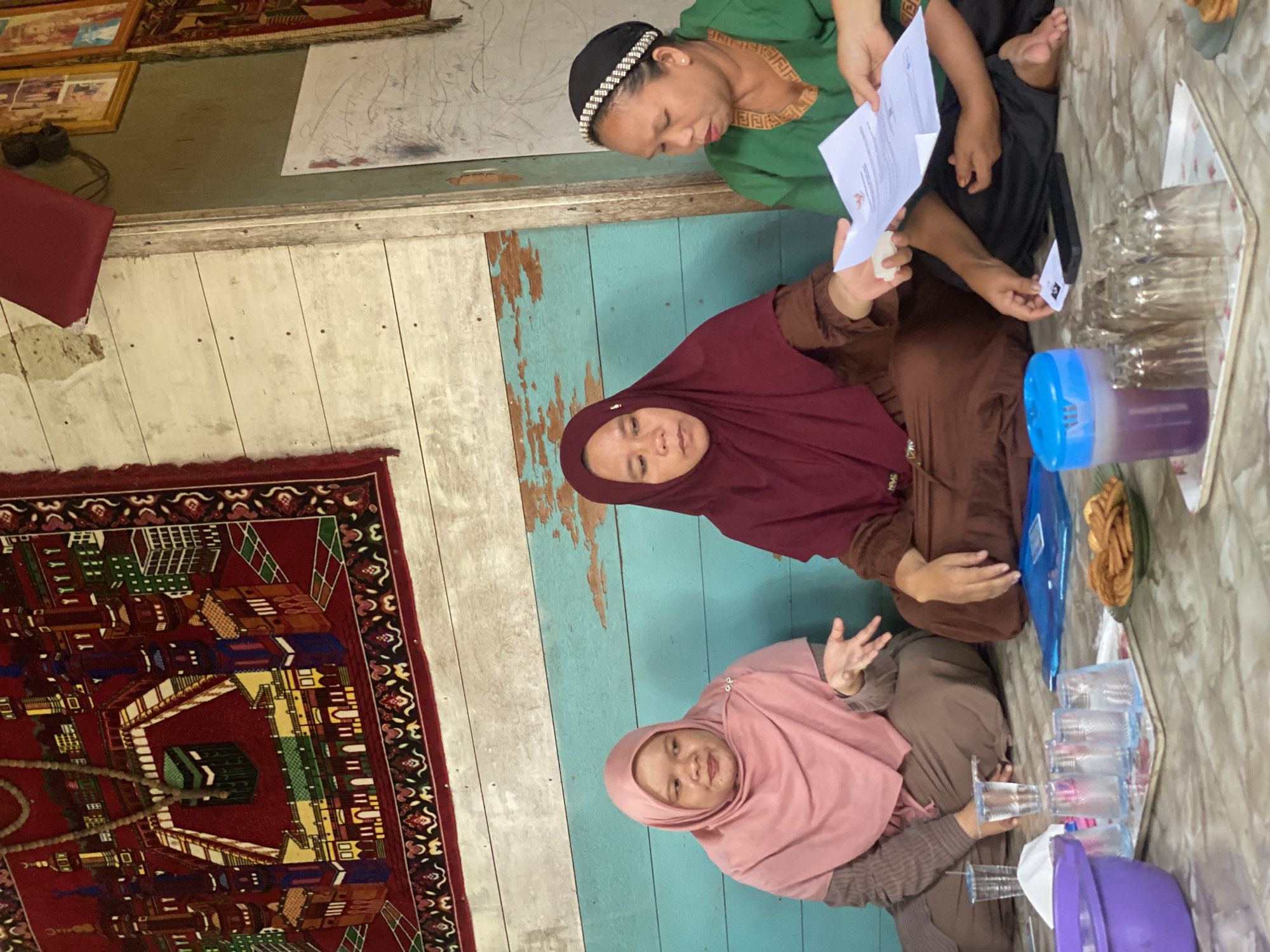 In an indoor setting that appears to be a simple wooden structure with turquoise painted walls and a wooden plank ceiling, three women are seated on the floor. The woman on the left is wearing a pink hijab and a brown sweater. The woman in the center wears a maroon hijab and a brown dress. The woman on the right is dressed in a green top and black pants, with a black head covering adorned with white beads. She is holding and presenting a document, possibly for review, to the woman in the maroon hijab, who is reaching out to take it. A light-colored rug with a detailed Islamic motif, depicting a mosque and its surroundings, hangs on the wall behind the women. In the foreground, on a tray, are several clear plastic cups, a purple bowl, a blue container with a purple liquid, and a small plate of cookies. The floor is tiled and appears to be the same surface as the walls. The scene suggests a gathering for a discussion or meeting, possibly within a modest dwelling in Bawahan Selan, Indonesia, given the context provided.